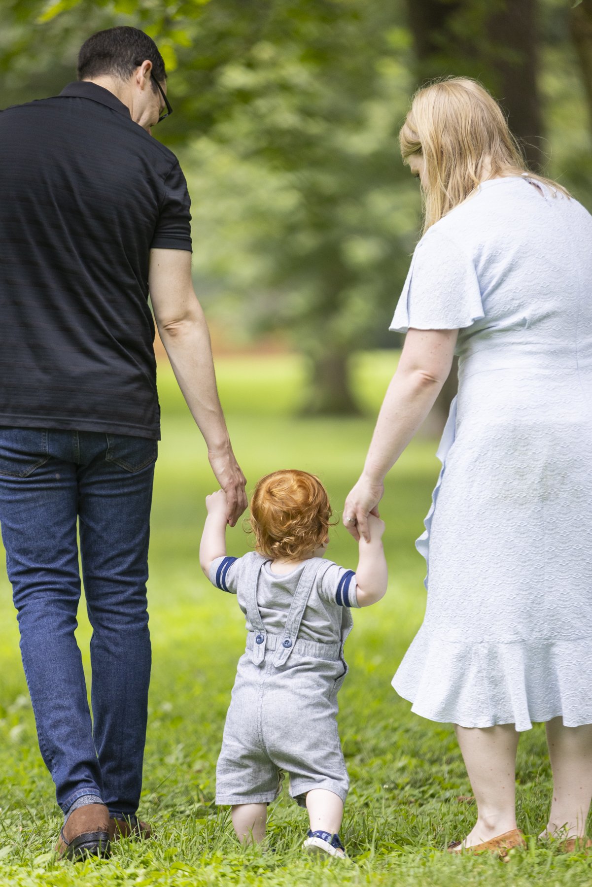 A toddler with red hair walking between a man and a woman in a park, holding their hands.