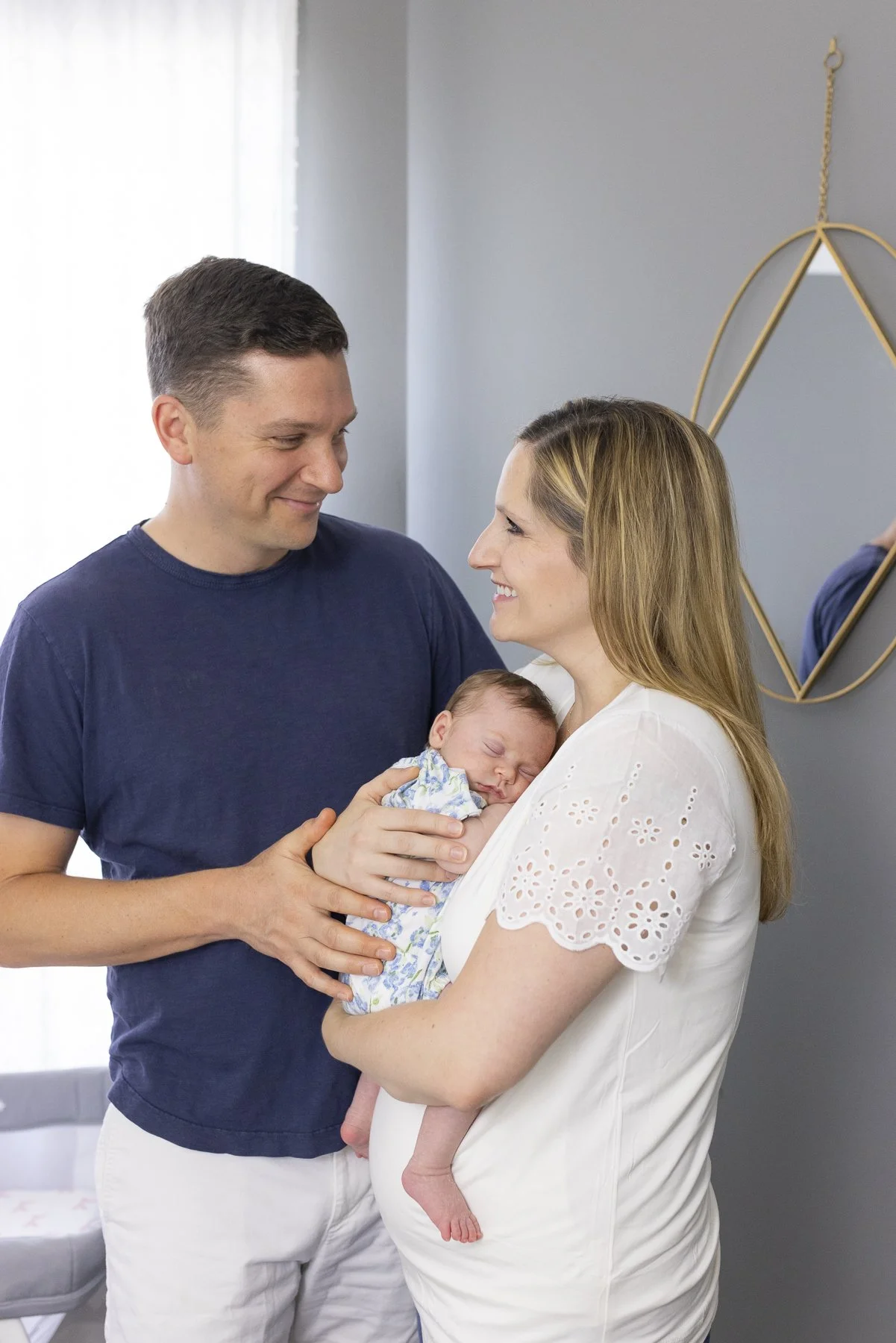 A couple holding a sleeping newborn baby, smiling at each other in a cozy room.
