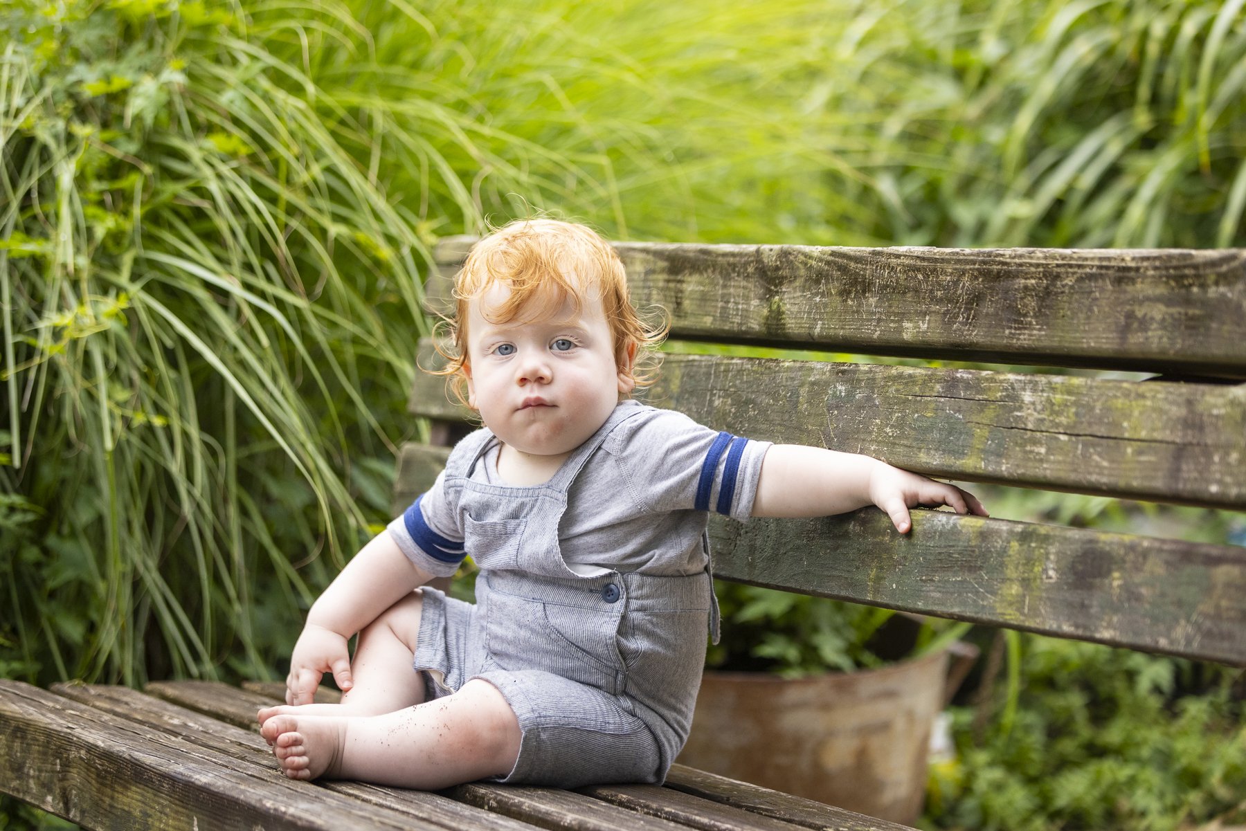 A young red-haired boy with blue eyes sitting on a wooden park bench surrounded by green plants, wearing a gray outfit with blue accents, looking directly at the camera.