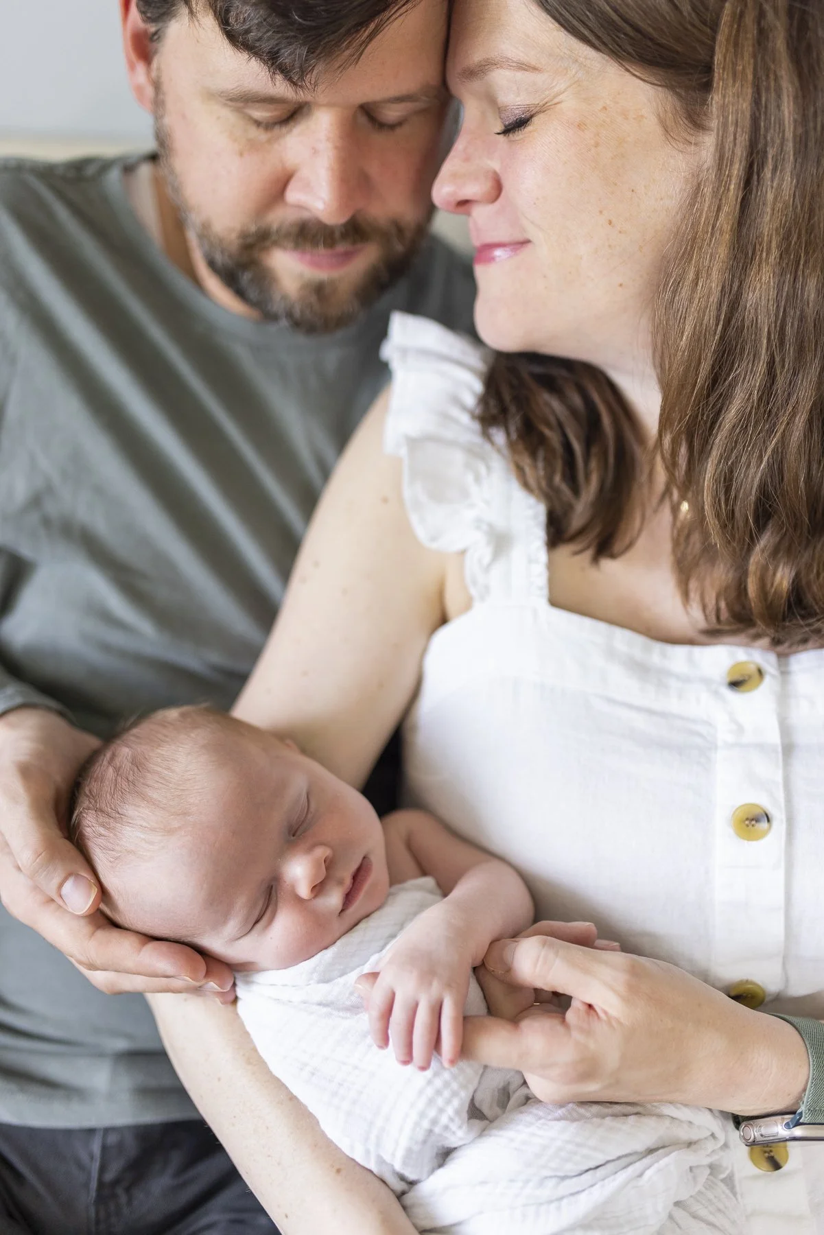 A happy family scene with a woman holding a sleeping baby, a man leaning in with his forehead touching the woman's, all appearing joyful and loving.