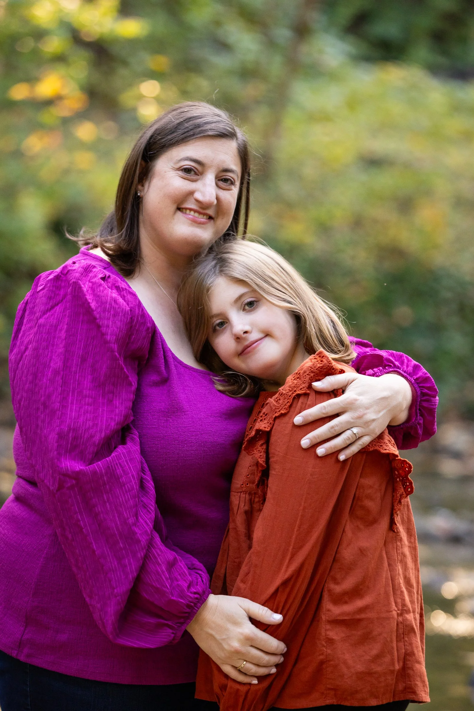 A woman and a young girl hugging outdoors with blurred trees in the background.