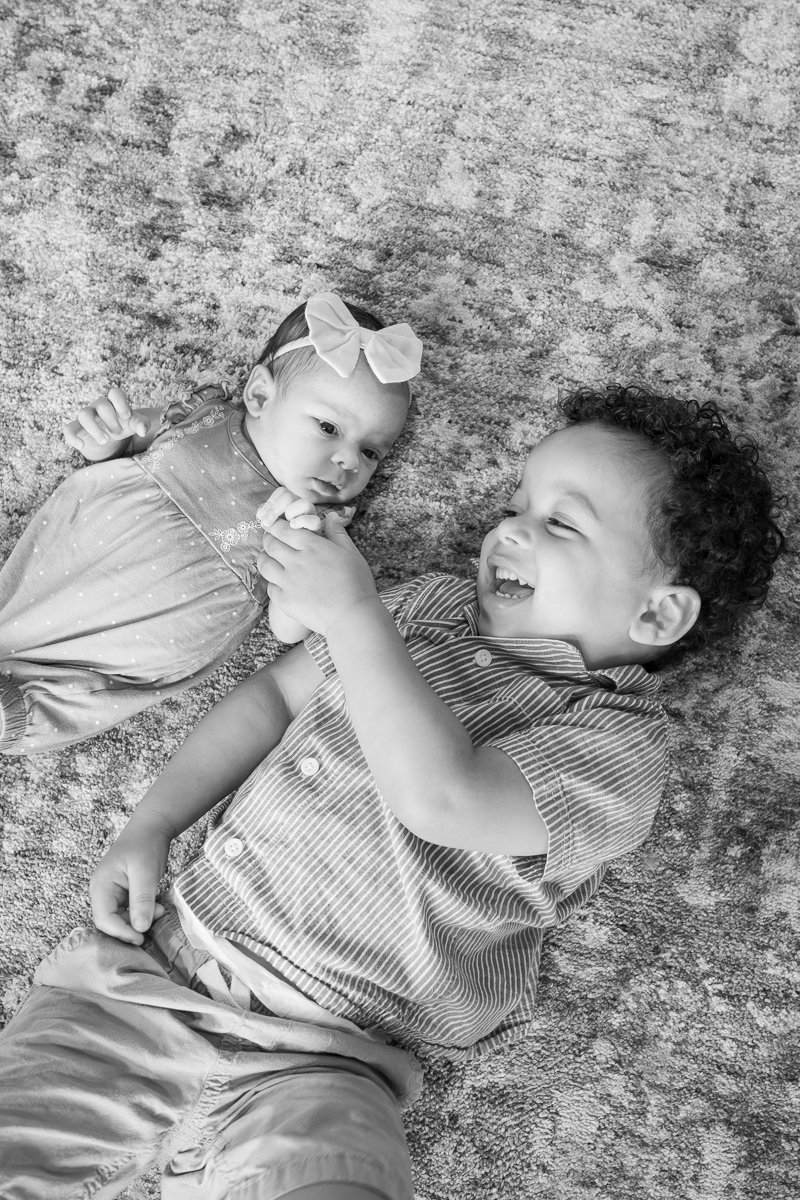 A young boy and girl lie on a carpet, holding hands, with the boy smiling and the girl looking at the boy.