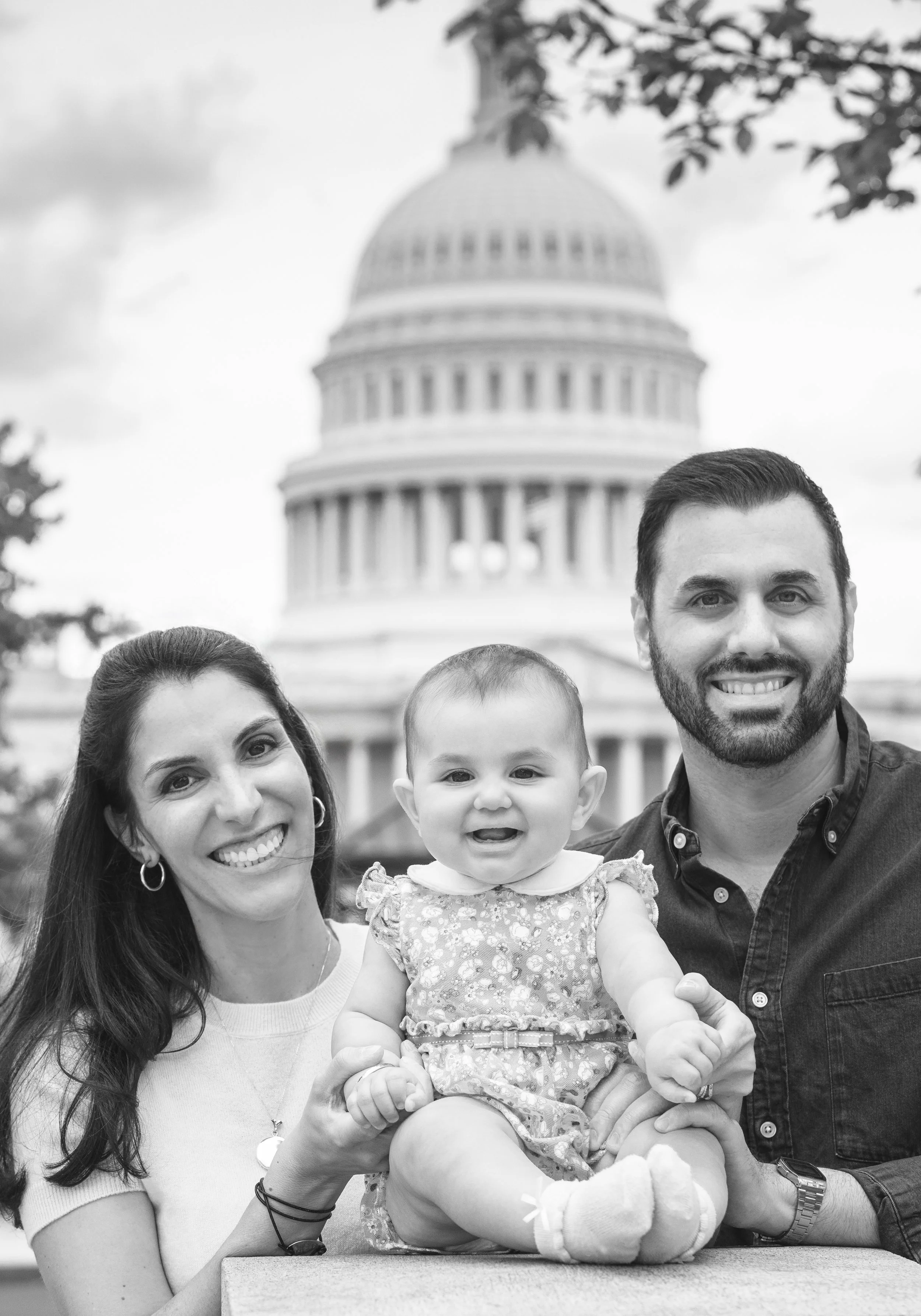 A family of three, a woman, a man, and a baby girl, smiling and posing together outdoors in front of the U.S. Capitol building.