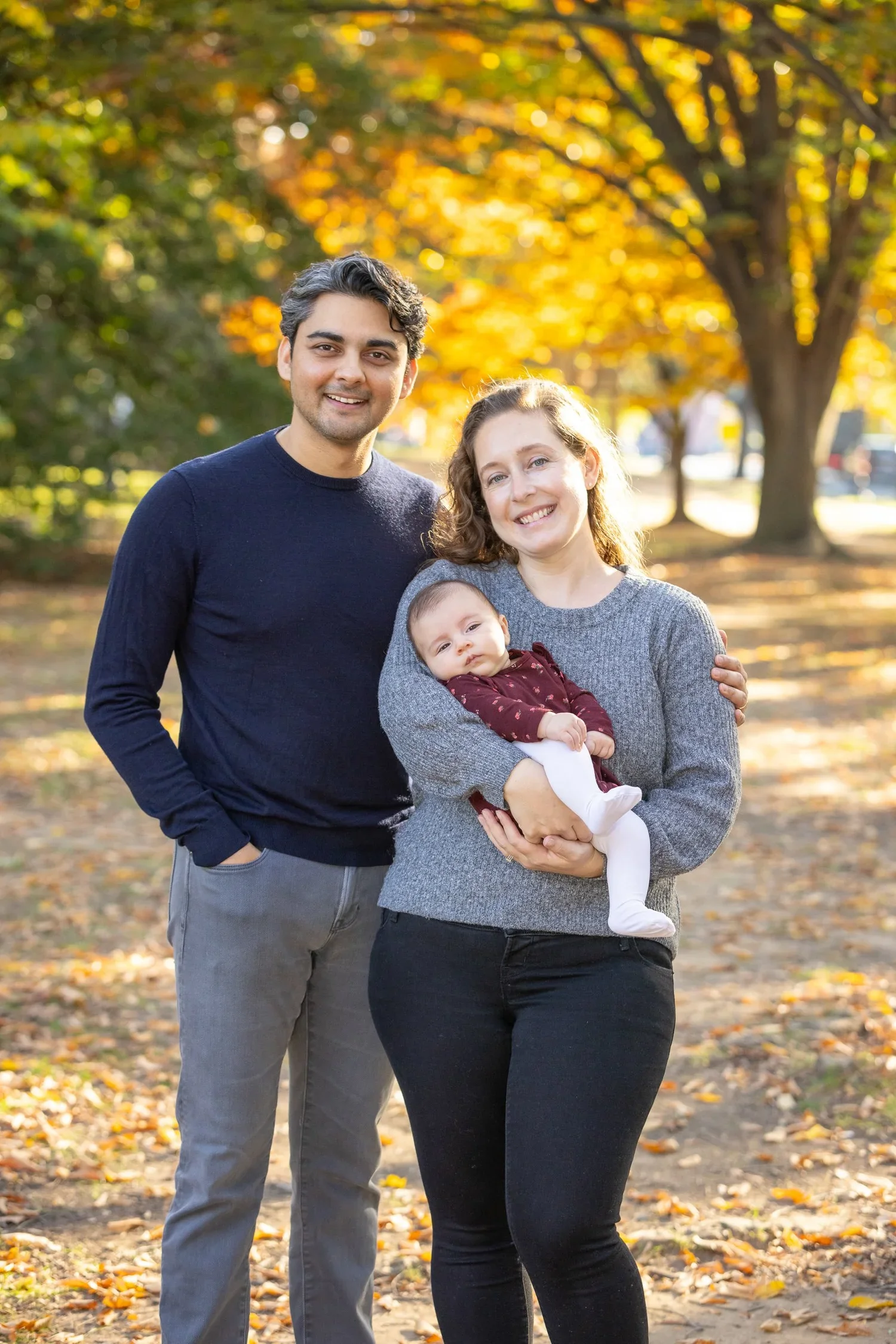 A family of three standing in a park during autumn, with colorful fall leaves on the ground and trees in the background. The woman is holding a baby, and a man is standing beside them, all smiling.
