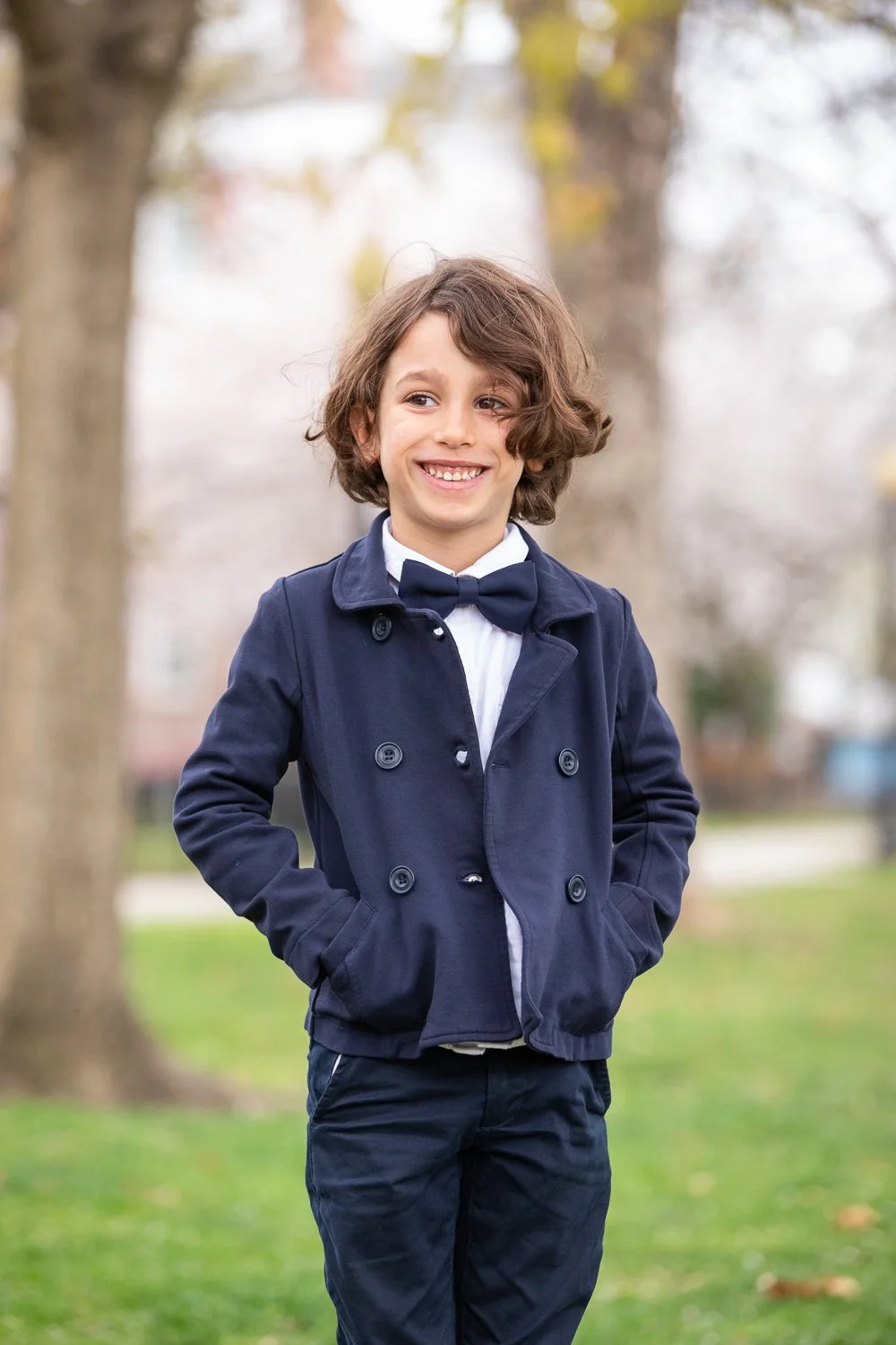 Young boy smiling outdoors in a navy blue coat, white shirt, and bow tie, standing in a park with trees and grass, autumn background.