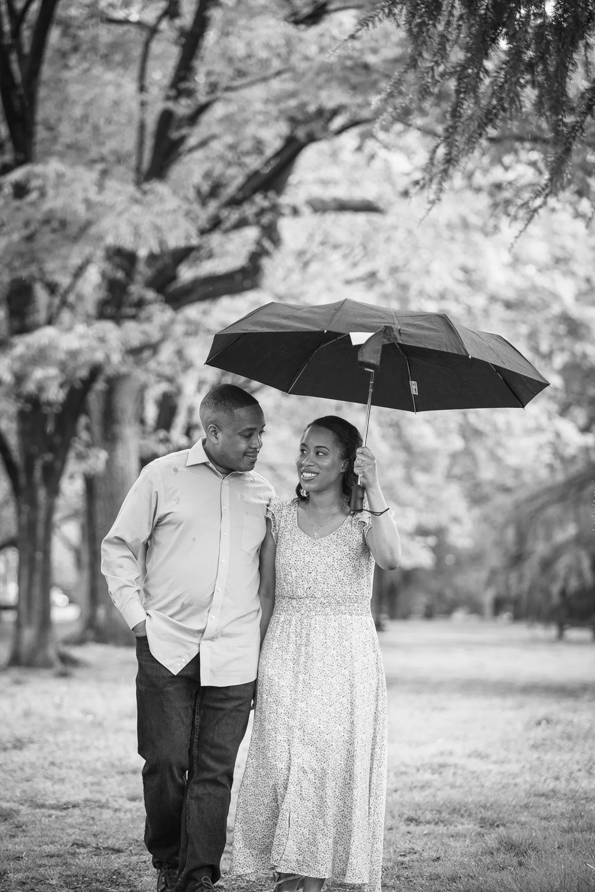 A smiling couple walks outside under an umbrella on a rainy day, surrounded by trees in a park.