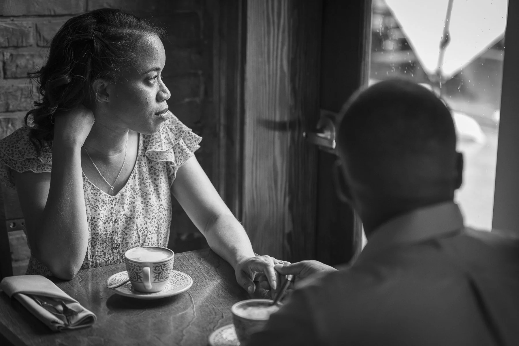 A woman with curly hair and wearing a patterned top holding hands with a man wearing glasses and a face mask, sitting at a table in a cafe or restaurant, with a coffee cup and a napkin on the table.