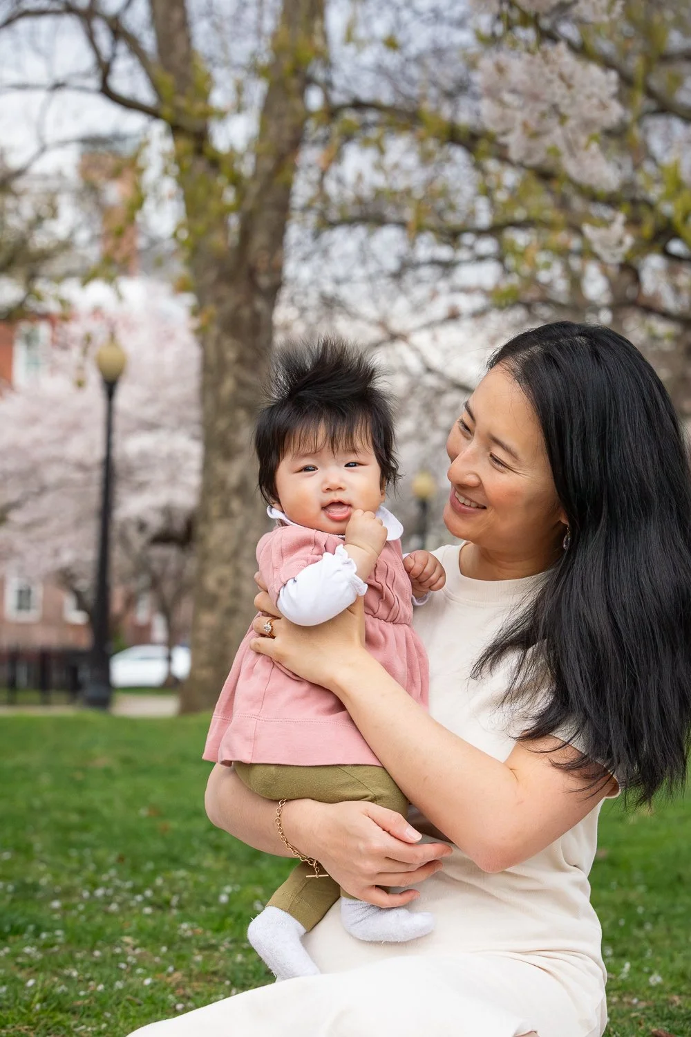 A woman holding a young child outdoors in a park with cherry blossoms in the background.