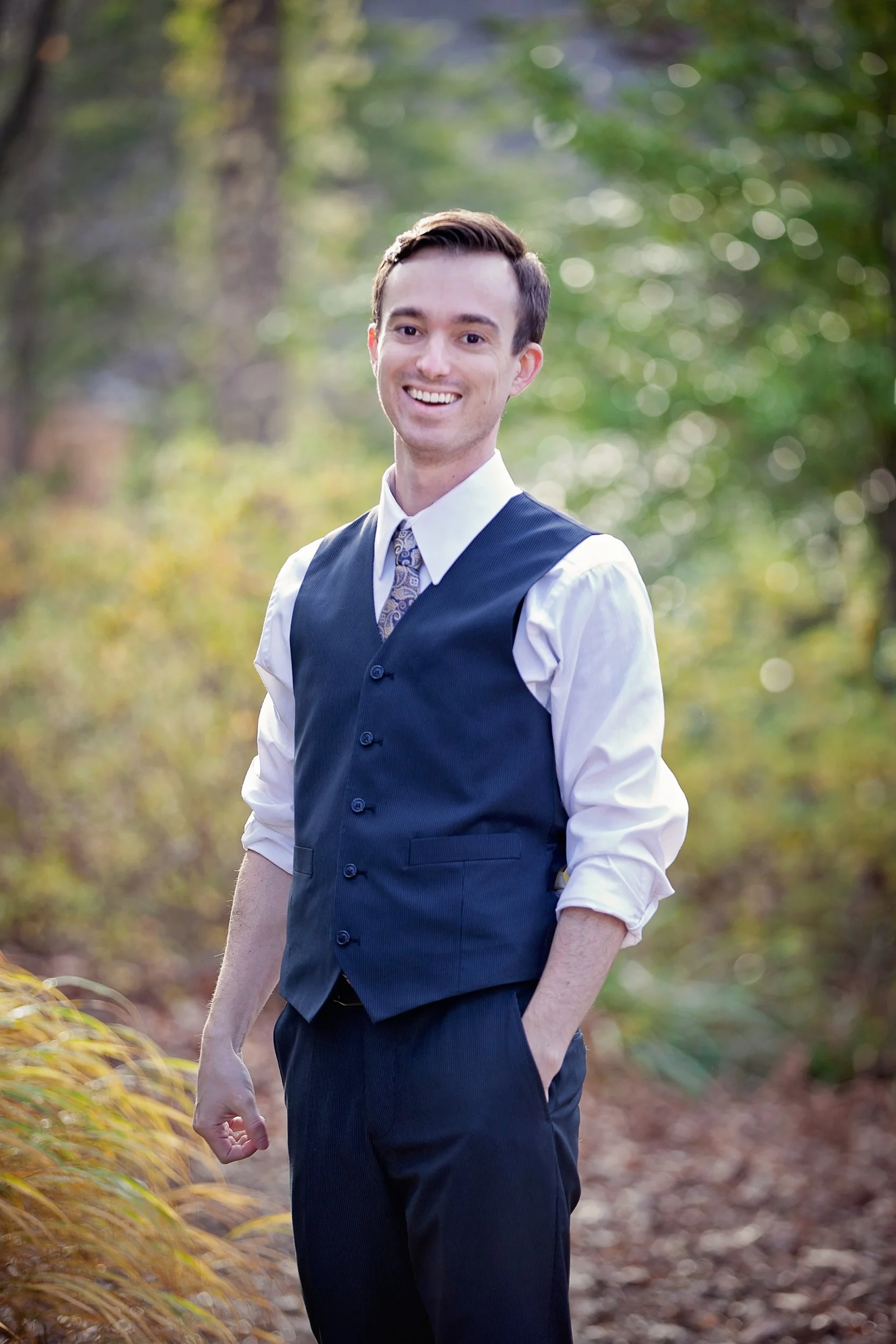 A man dressed in formal attire, wearing a vest over a white shirt and a tie, standing outdoors in a wooded area, smiling at the camera.