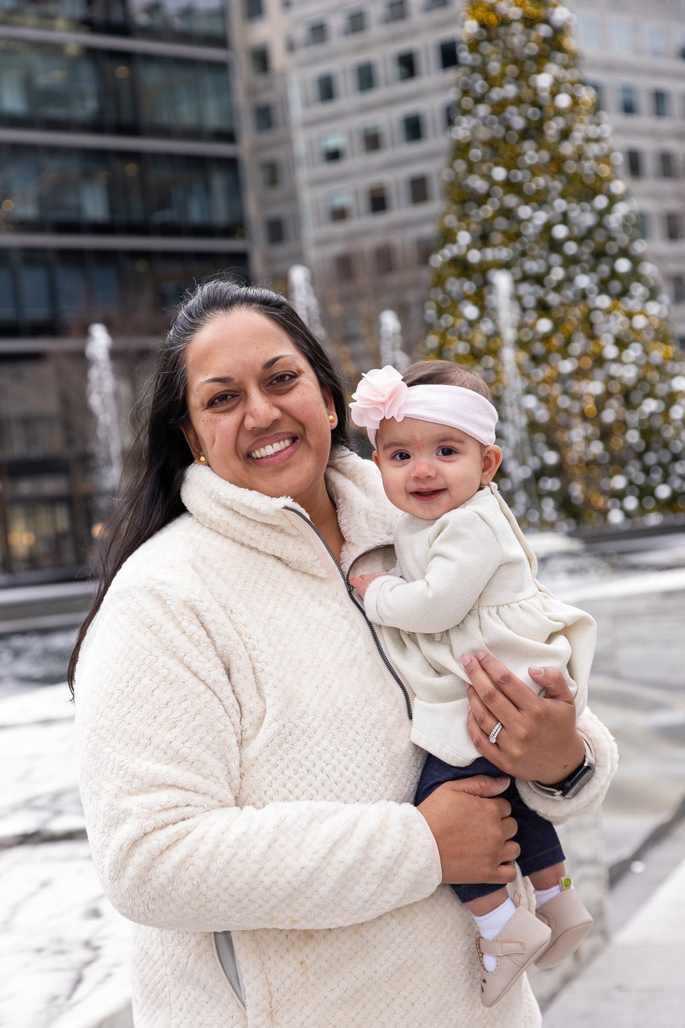 A smiling woman holding a young girl in front of a cityscape with a decorated Christmas tree and a fountain.