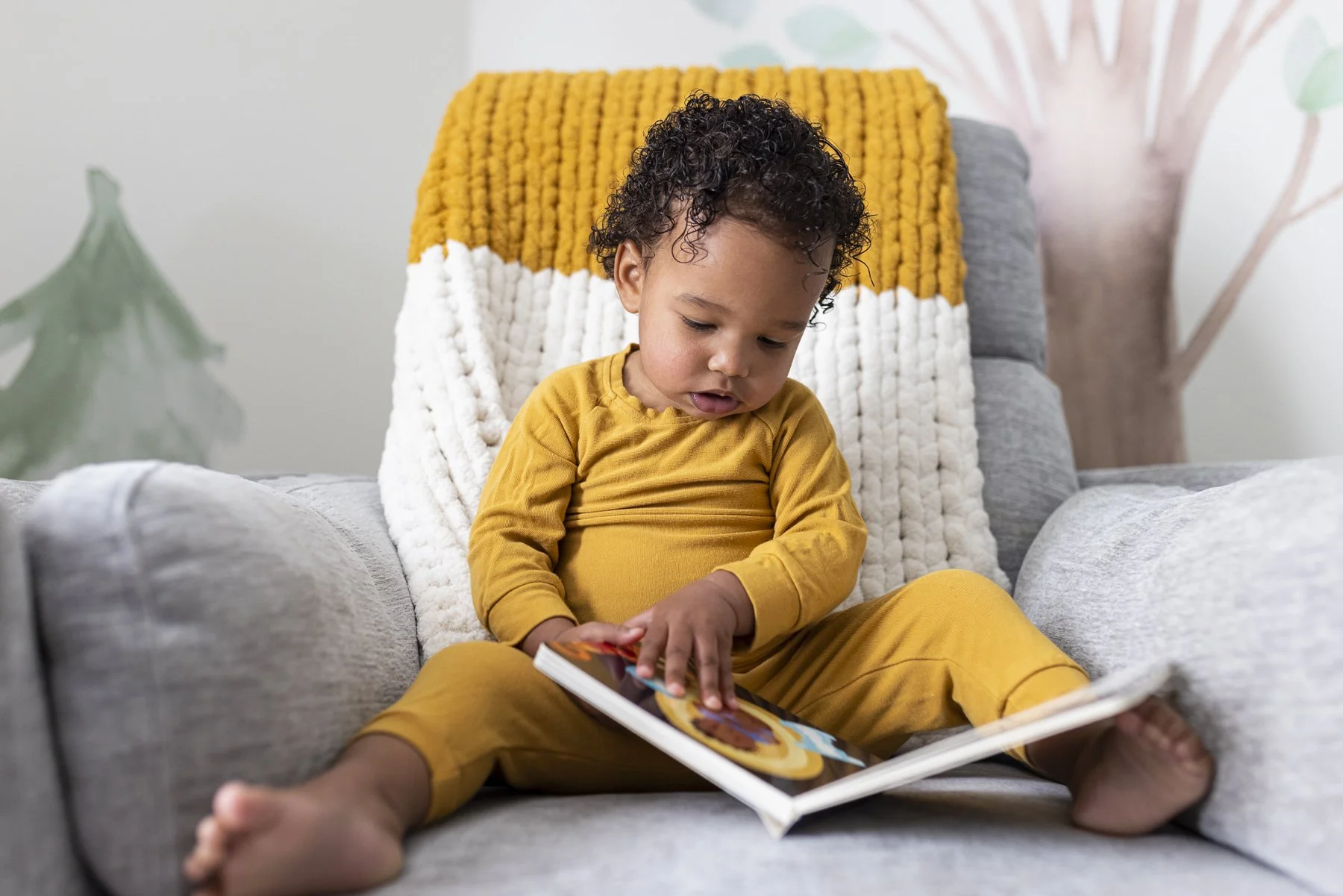 Young child sitting on a gray couch, wearing matching yellow pajamas, looking at a colorful children's book.