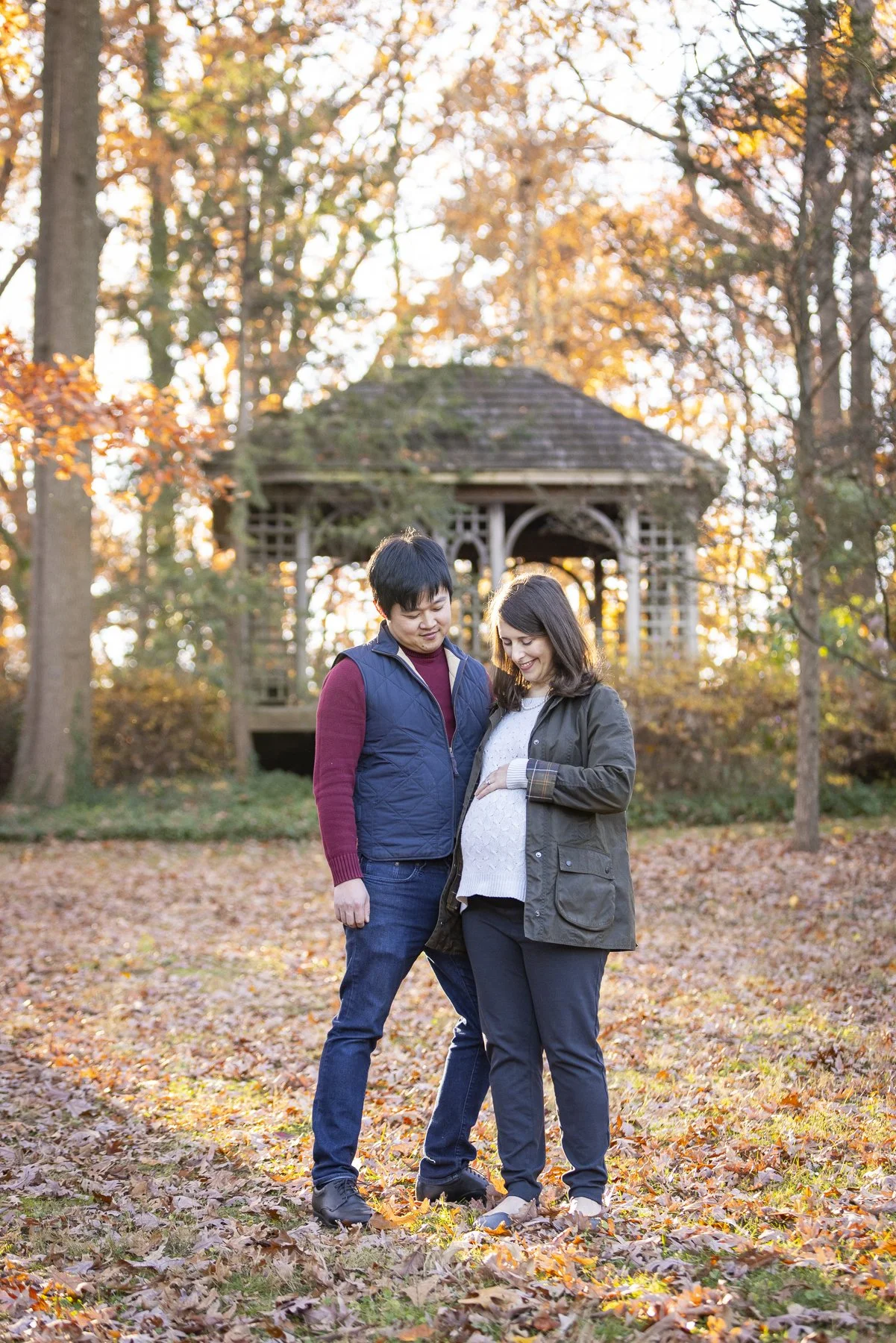 A couple, with the pregnant woman holding her belly, standing closely together in a park during autumn with fallen leaves and a gazebo in the background.