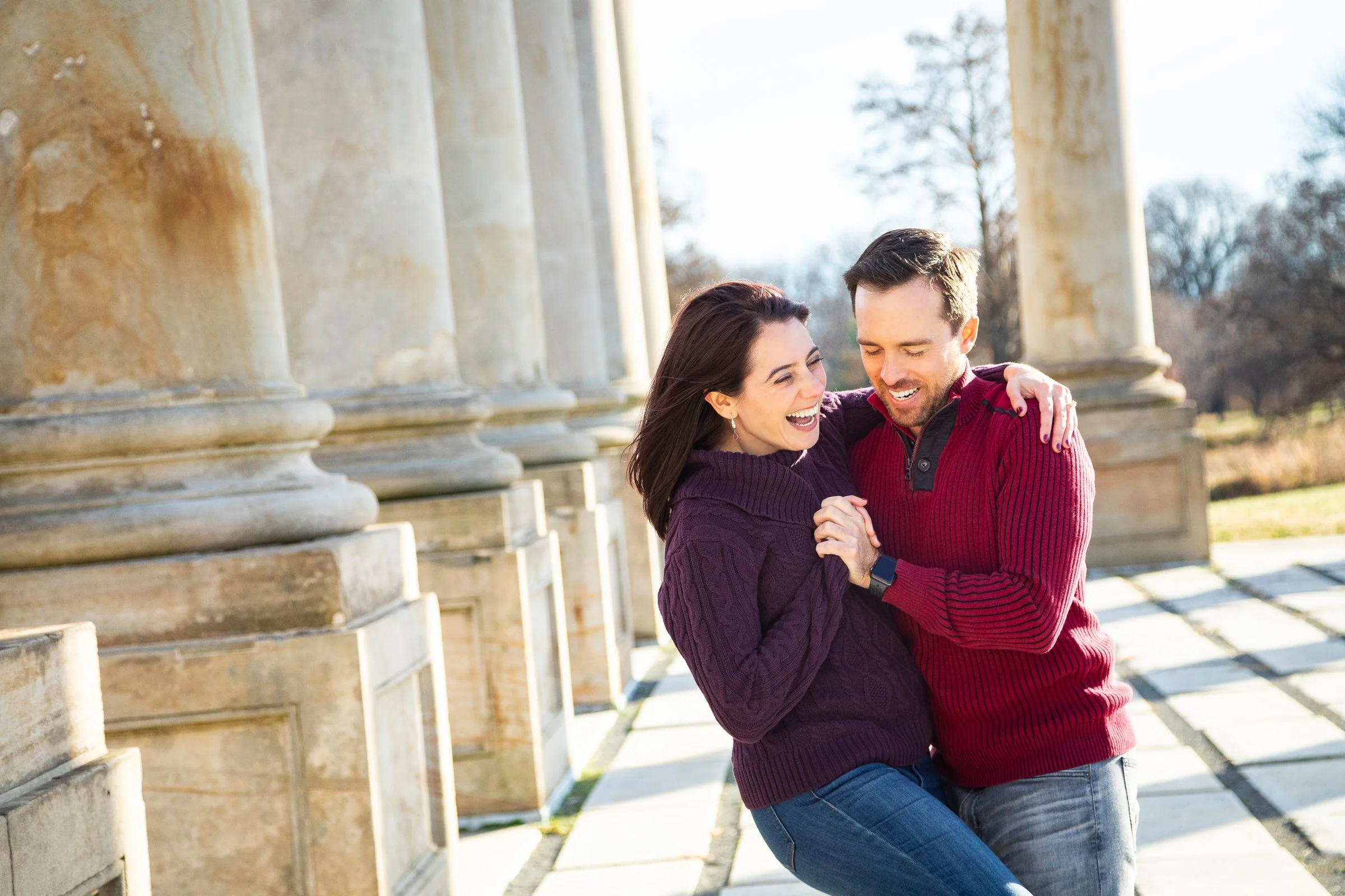 A smiling man and woman enjoying an outdoor moment, standing close together with the woman's arms around the man's shoulders, in front of historic stone columns.