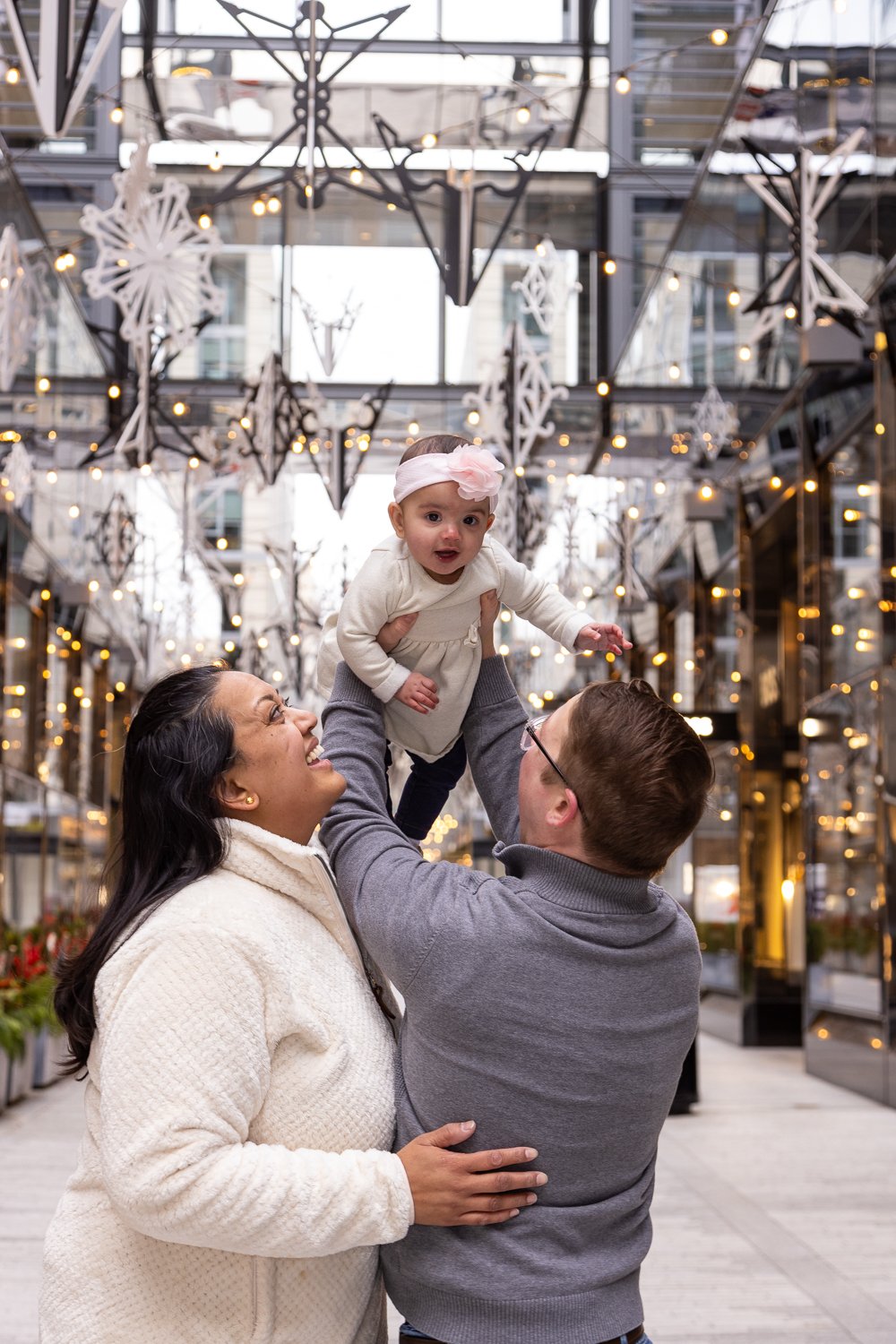 A family playing together under festive holiday decorations with snowflake and star-shaped lights hanging overhead in an urban shopping area.