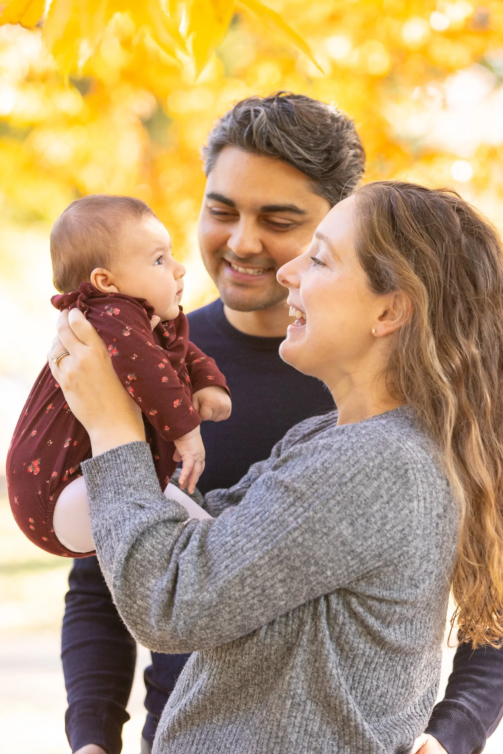 A family of three outdoors in autumn, with trees in the background, smiling and looking at each other. The mother holds a baby girl dressed in a maroon outfit, and the father stands behind them.