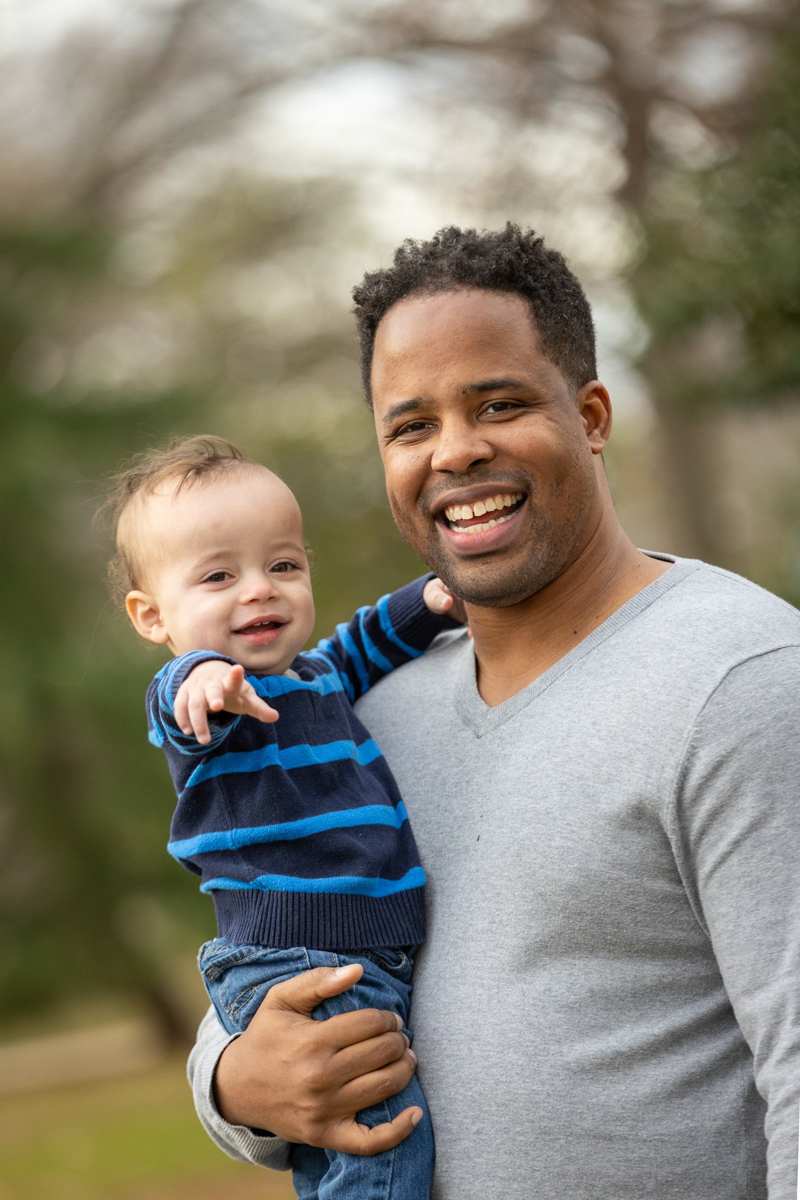 A man holding a smiling toddler outside in a park with trees in the background.