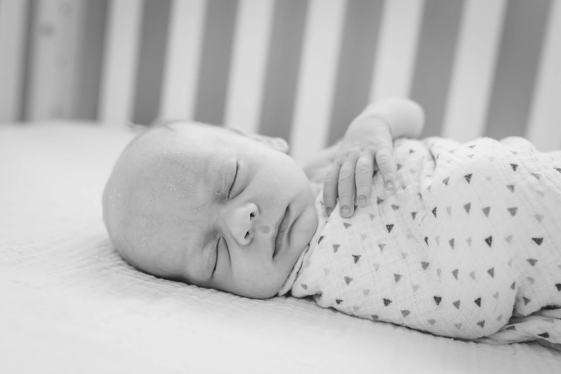 Black and white photo of sleeping baby lying on back with hand on chest, in a crib.