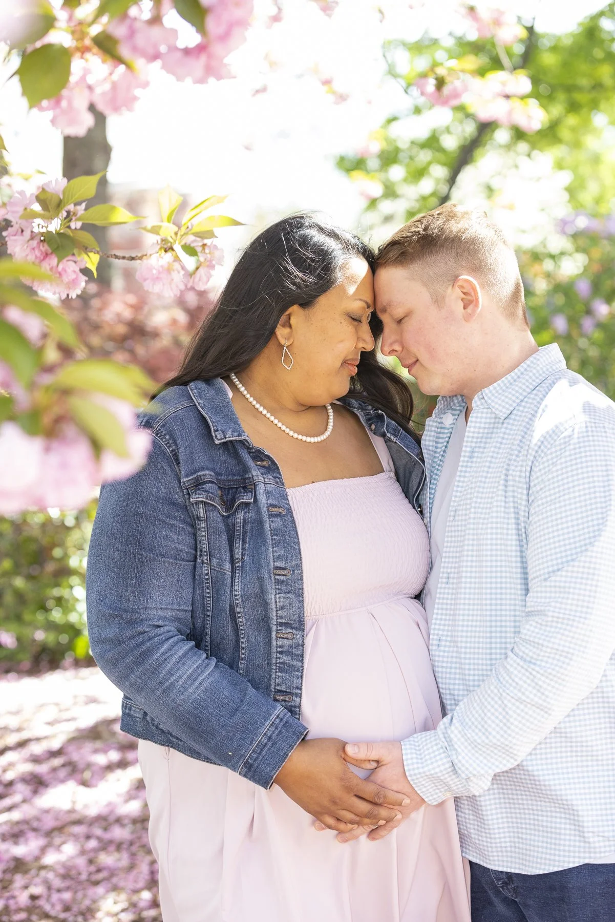 A couple with their foreheads and noses touching, gently holding hands, standing among pink flowering trees in a park on a sunny day.