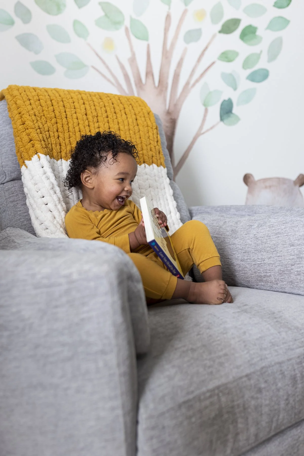 A young child with curly hair sitting on a gray sofa, laughing and holding a book, with a plush white and yellow blanket behind their back and a painted tree on the wall in the background.