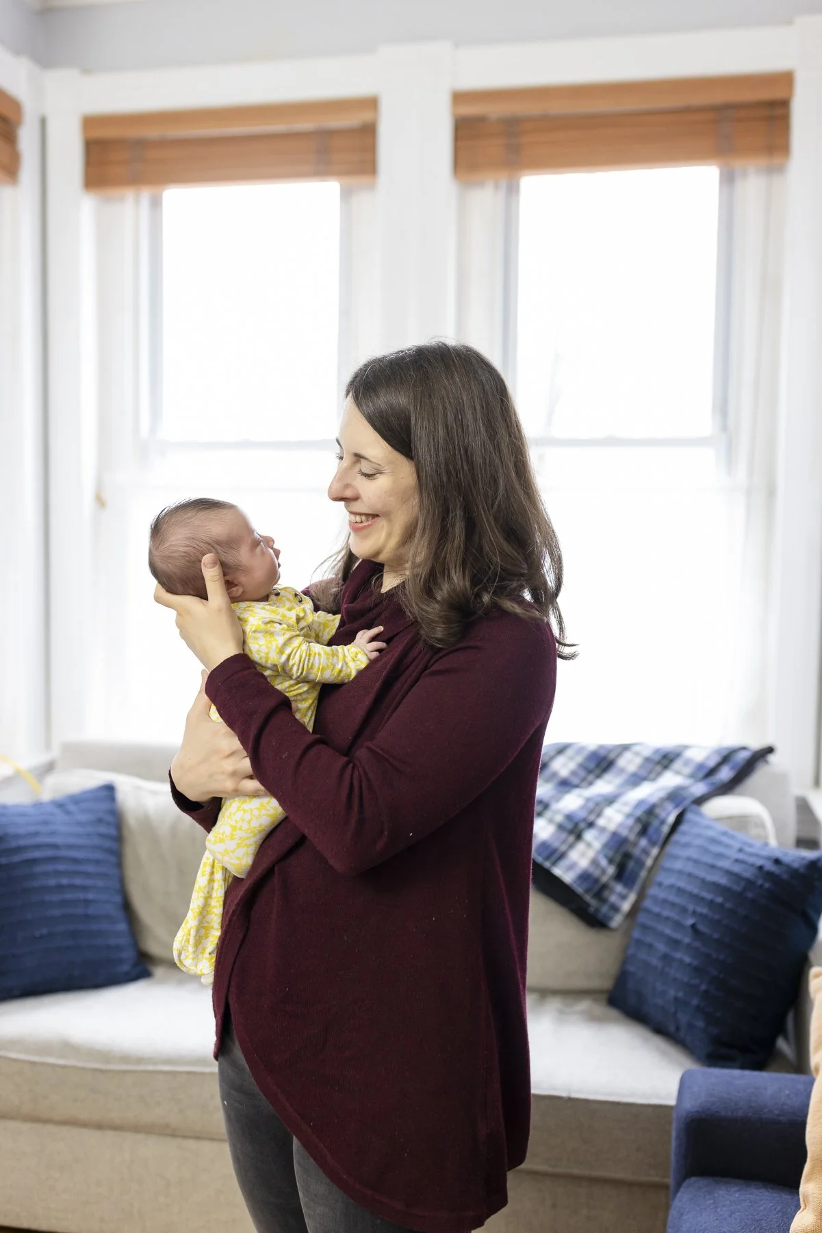 A woman holding a newborn baby inside a living room with a sofa and large windows in the background.