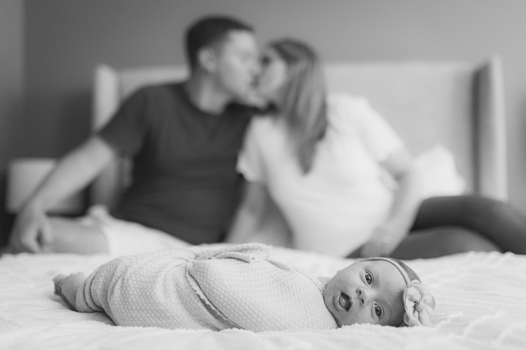 A baby lying on a bed with a pacifier clip and a flower headband, looking at the camera, while a couple in the background kisses in a bedroom setting.