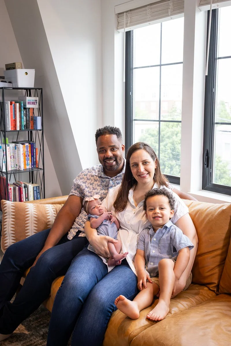 A family of four sitting on a tan leather couch in a bright living room, with large windows and a bookshelf in the background. The mother is holding a newborn, and a young boy is sitting beside her, smiling.