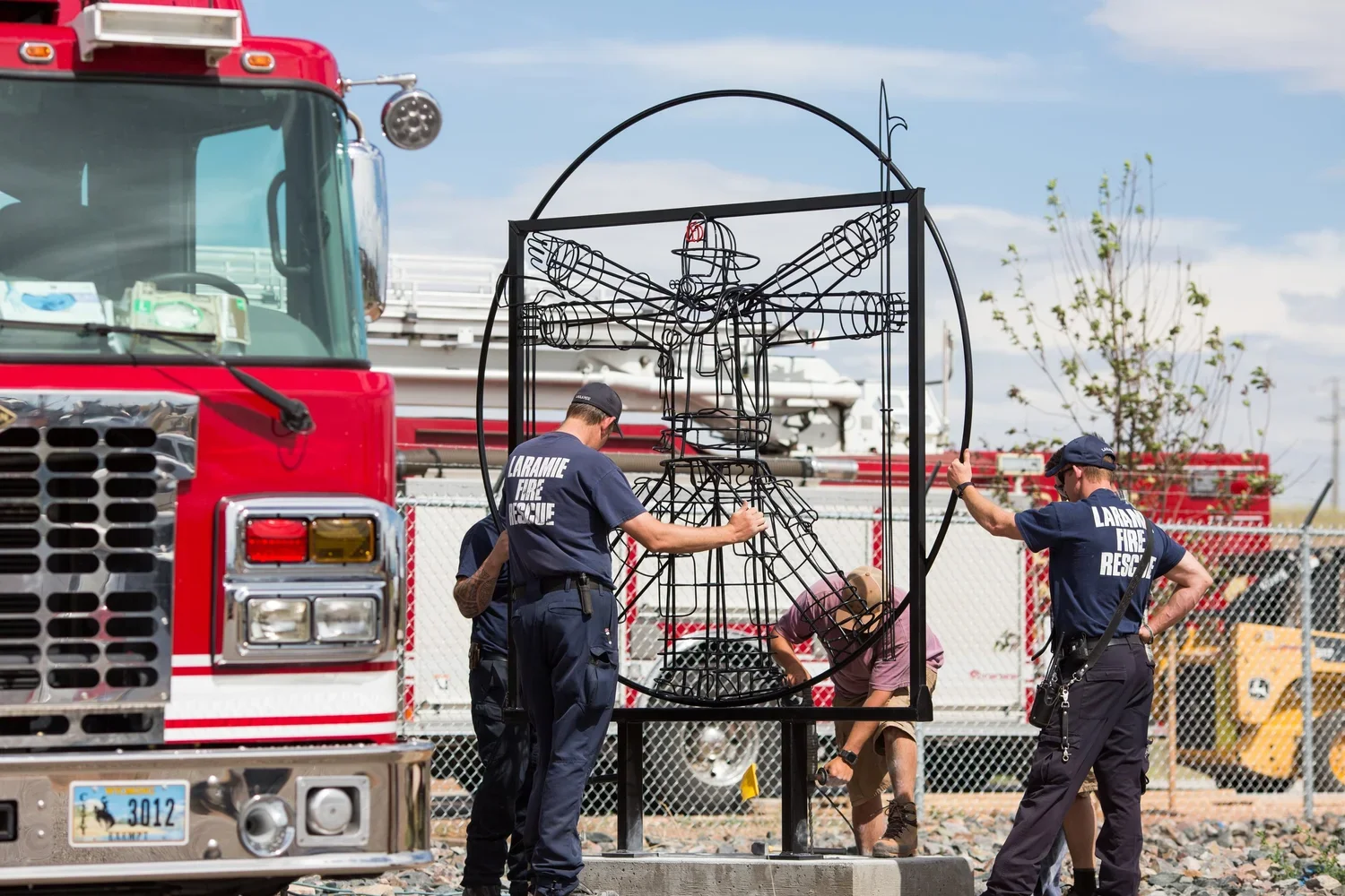 Red fire truck in right corner with fire fighters gathered around, assisting artist install their work