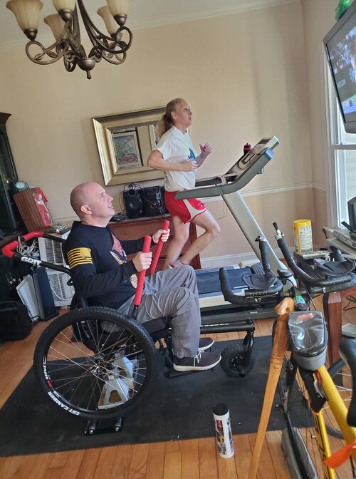 Rider pushes GRIT Freedom Chair mounted indoors next to adult running on treadmill