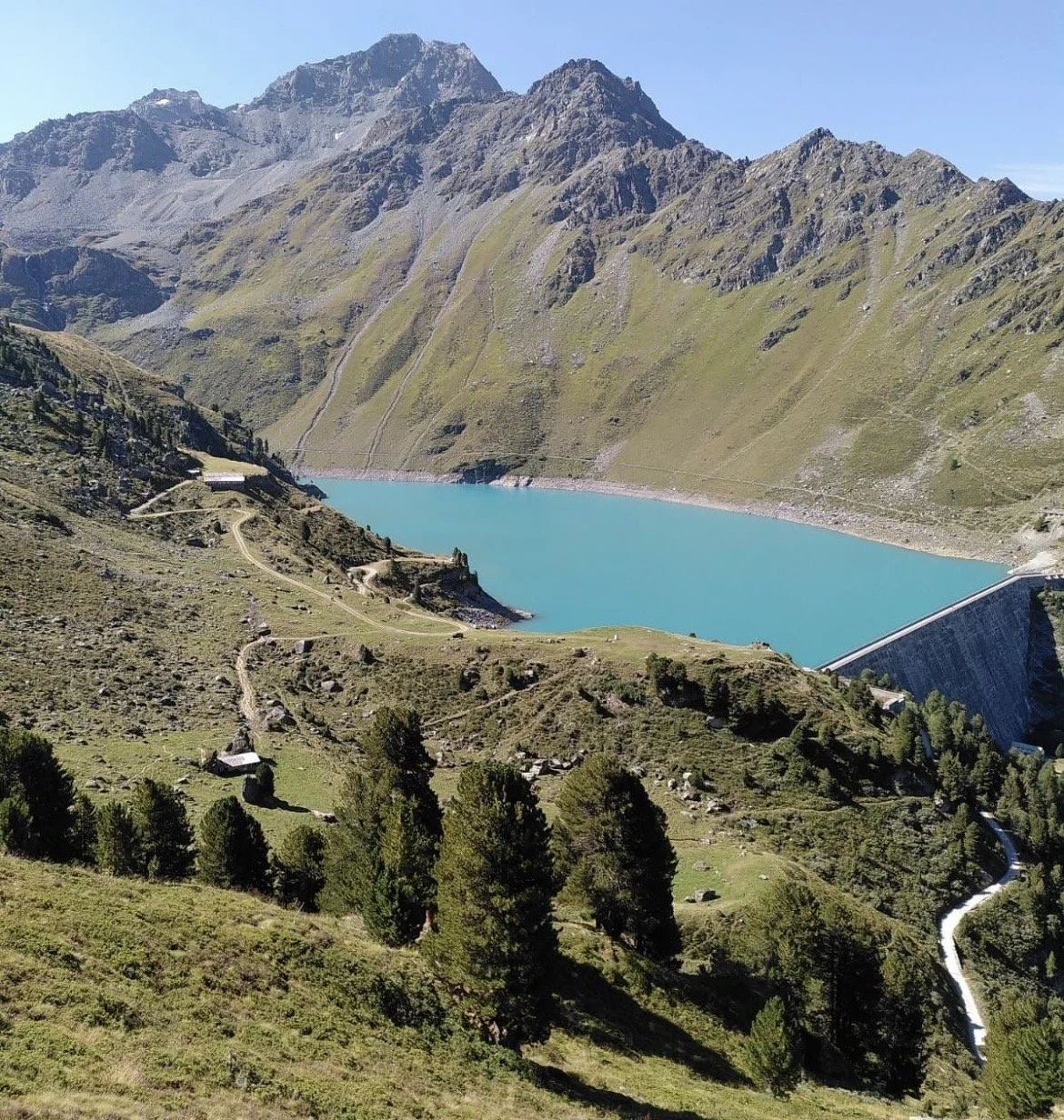 Lac de montagne entouré de collines verdoyantes et de montagnes escarpées sous un ciel dégagé, with un petit barrage visible sur le côté droit.