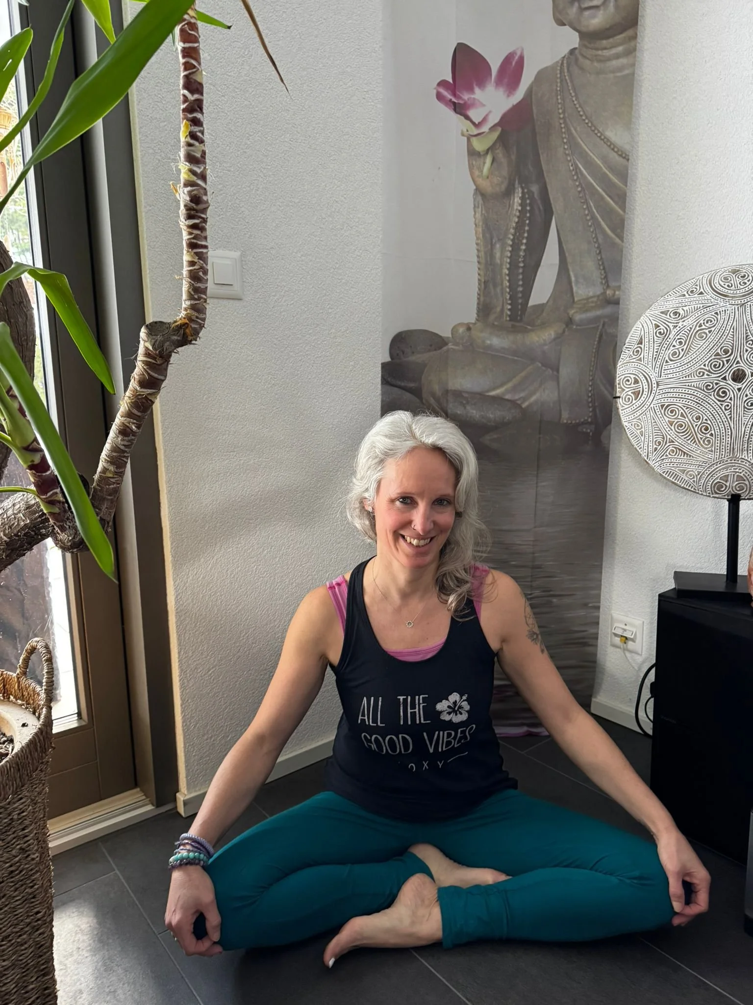 Femme assise sur le sol en position de lotus avec un sourire, portant un t-shirt noir avec l'inscription 'All the good vibes' et un pantalon vert-bleu, dans une pièce décorée de plantes et d'une œuvre d'art murale à portrait, près d'une grande fenêtre.