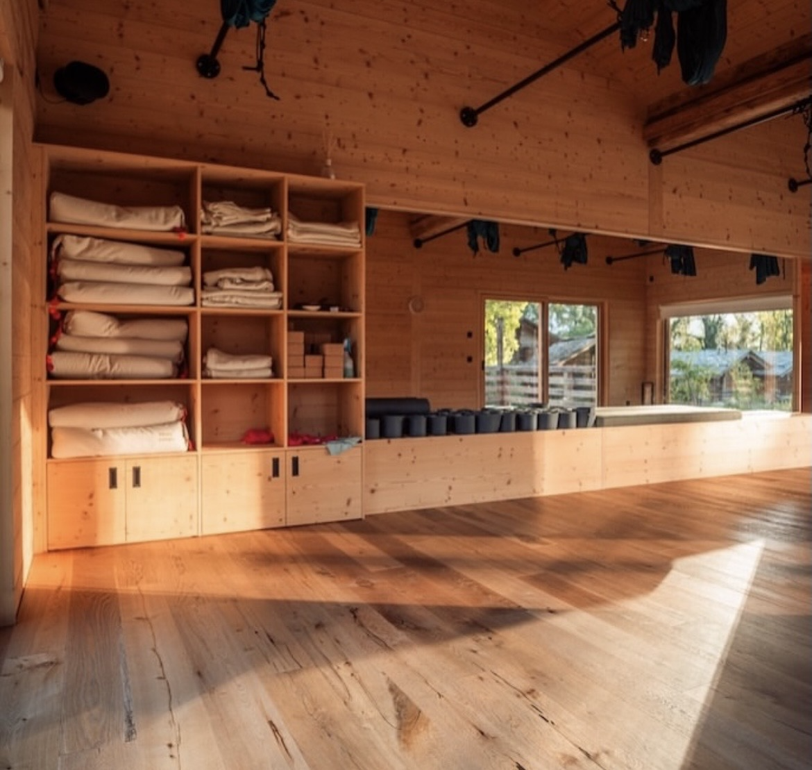 Salle de yoga ou entraînement avec étagère en bois contenant des tapis de yoga pliés et des blocs, fenêtre laissant entrer la lumière naturelle, et barres pour suspension suspendues au plafond.