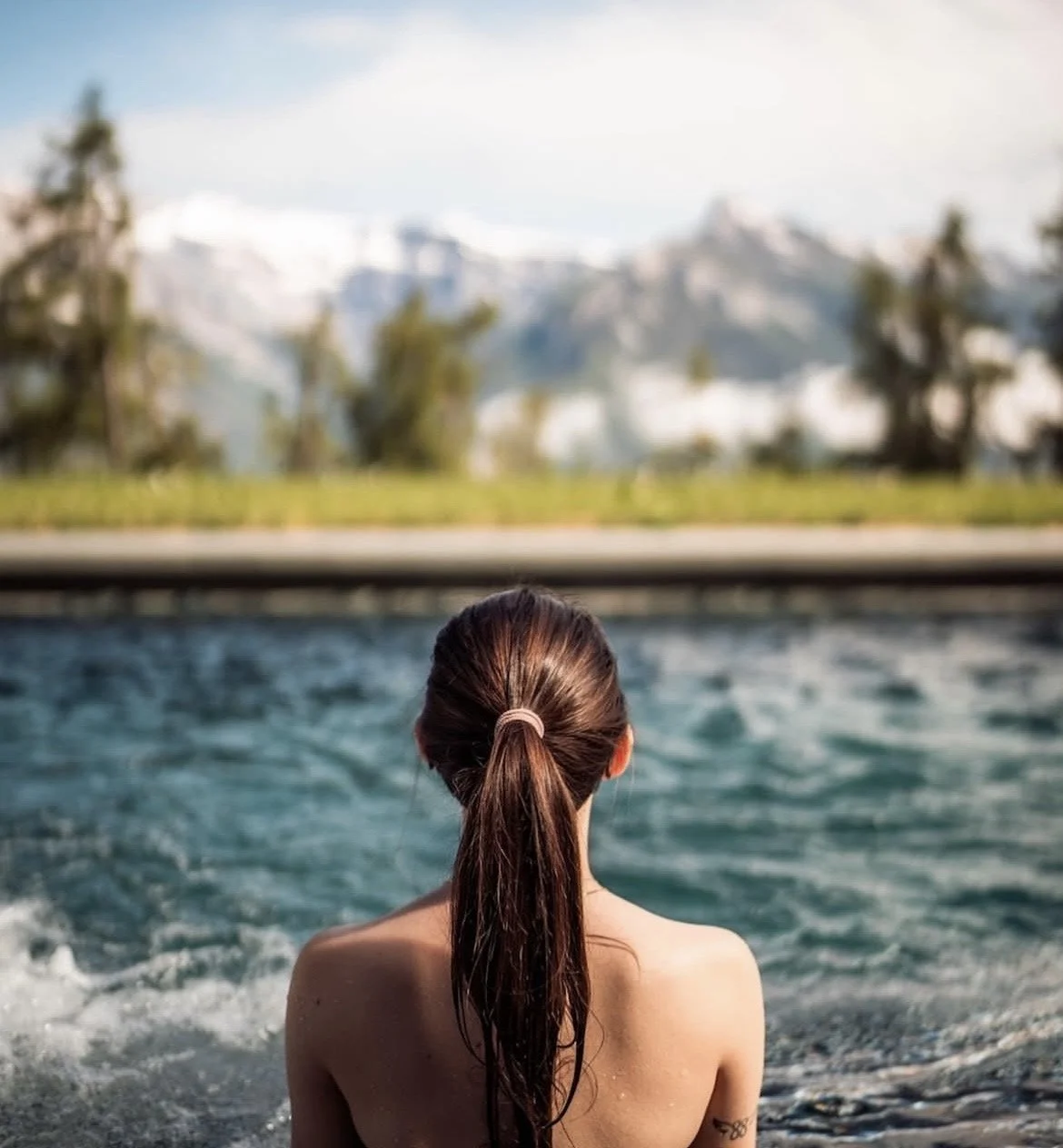 Femme avec cheveux longs attachés regardant un paysage montagneux depuis un jacuzzi ou une piscine.