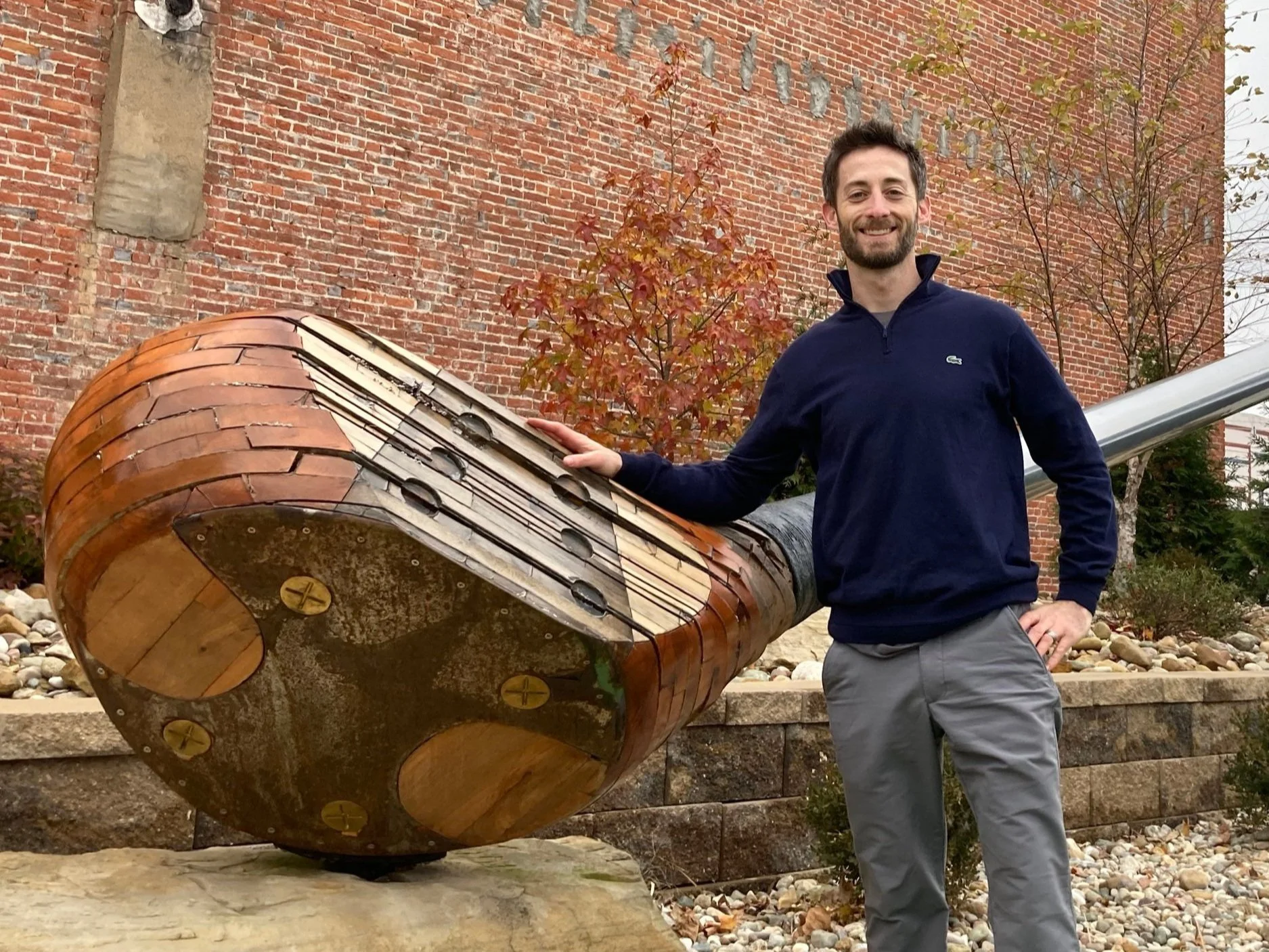 A smiling man with a beard, wearing a navy blue jacket and gray pants, stands next to a large decorative sculpture of a golf club, outdoors in front of a brick wall and autumn trees.