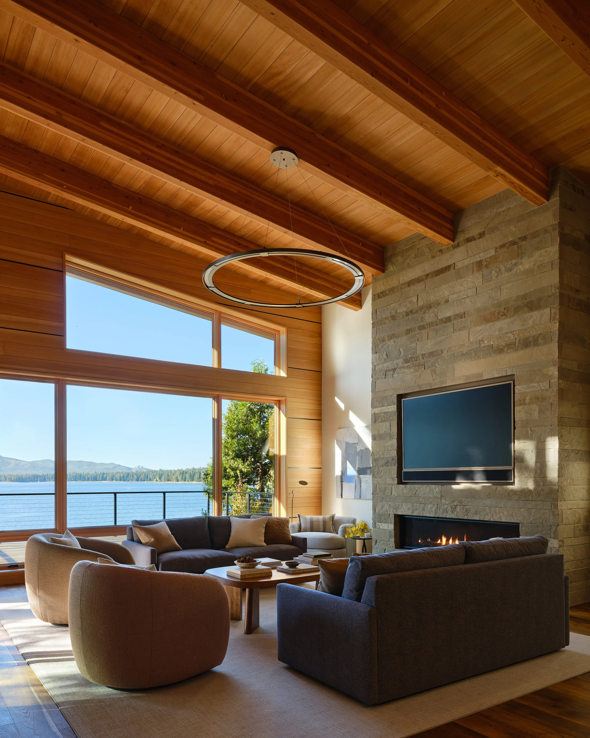 Living room with large windows, wooden ceiling, stone fireplace with television, and modern furniture overlooking water and trees outside.