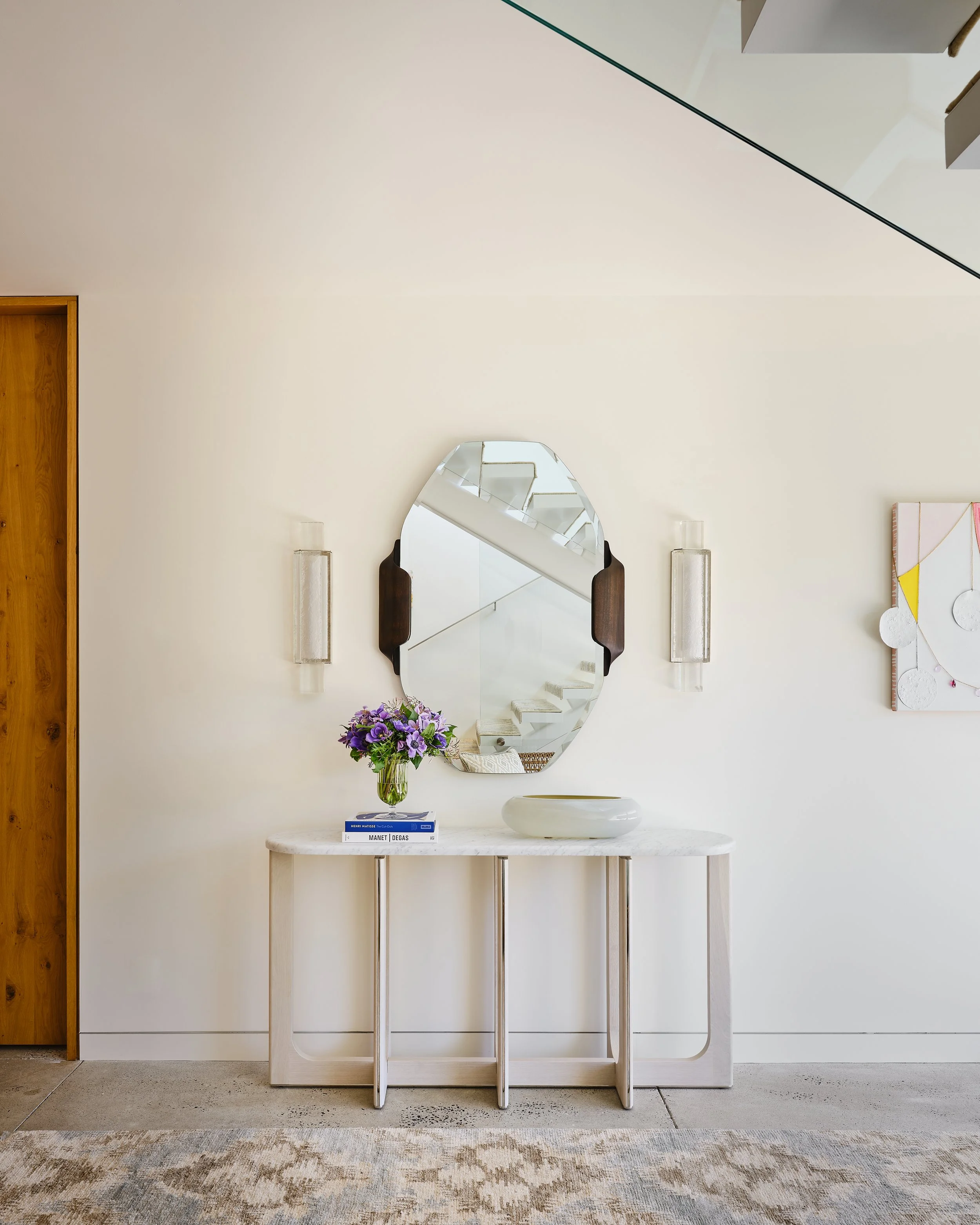 Modern entryway with a white table, a mirror, a flower vase, and books, against a plain wall with wall sconces and a colorful artwork.