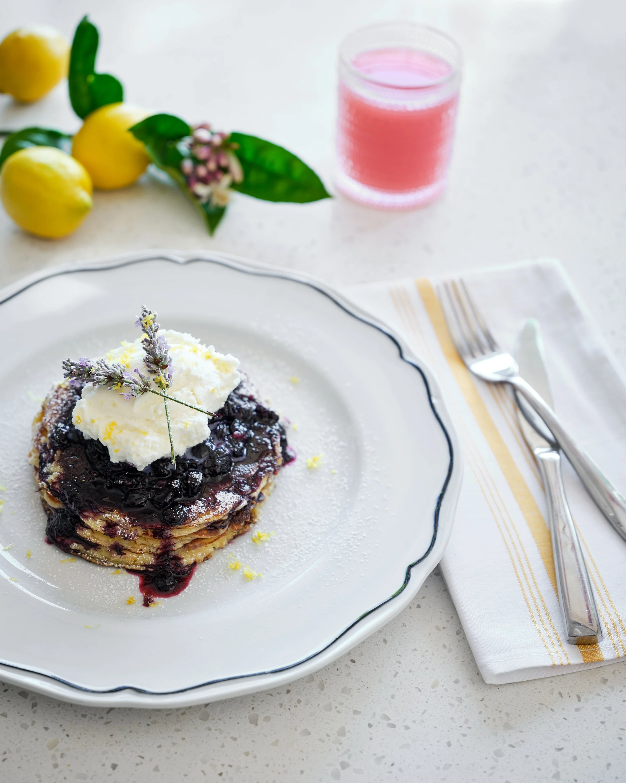 Stack of pancakes topped with whipped cream and blueberry syrup on a white decorative plate, with a sprig of lavender on top, on a white table with a fork, knife, and napkin, alongside lemons, a leaf, and a glass of pink beverage in the background.