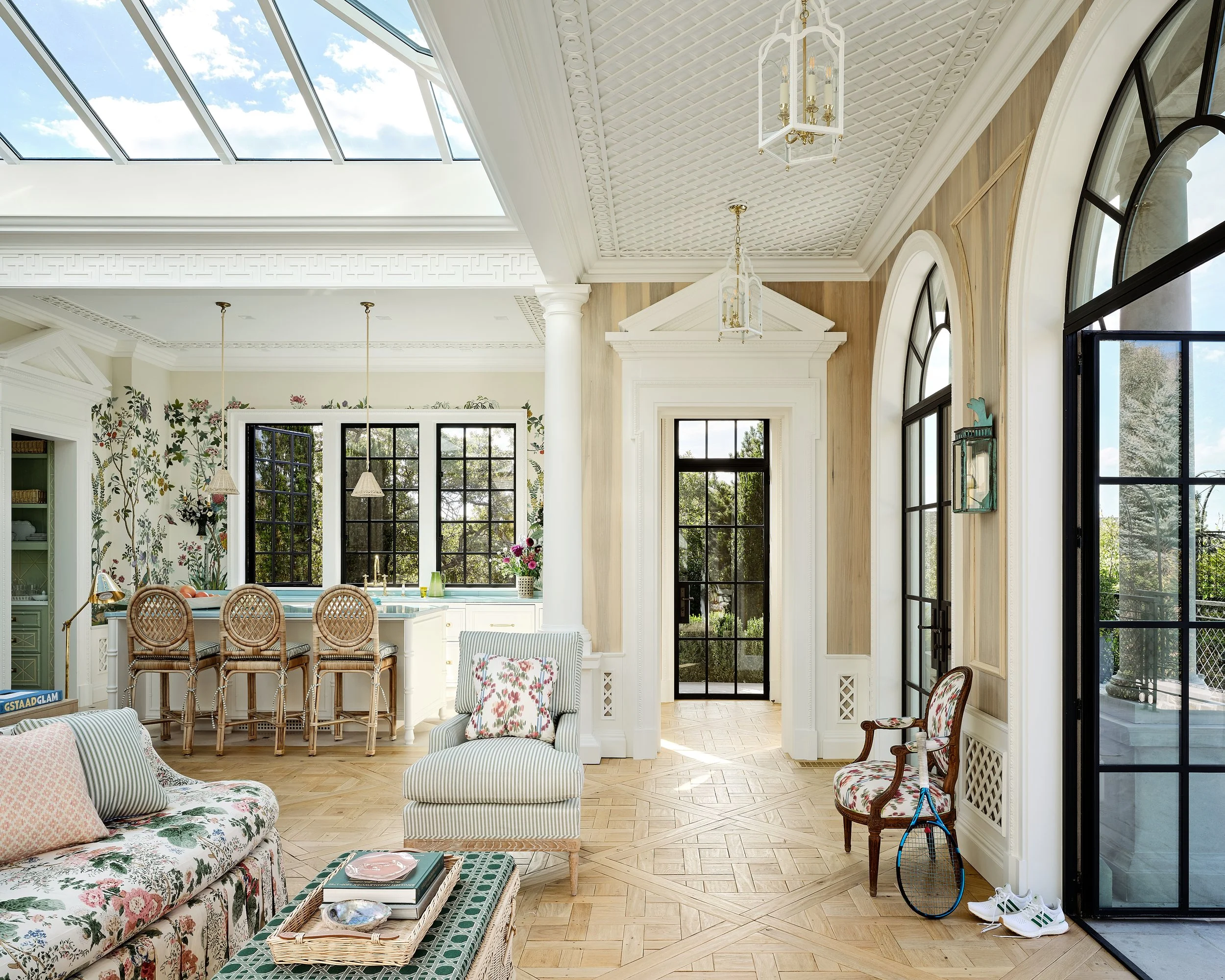Spacious, brightly lit living area with large windows and skylight, decorated in a cozy, floral style with white, green, and pastel accents, and a kitchen in the background.
