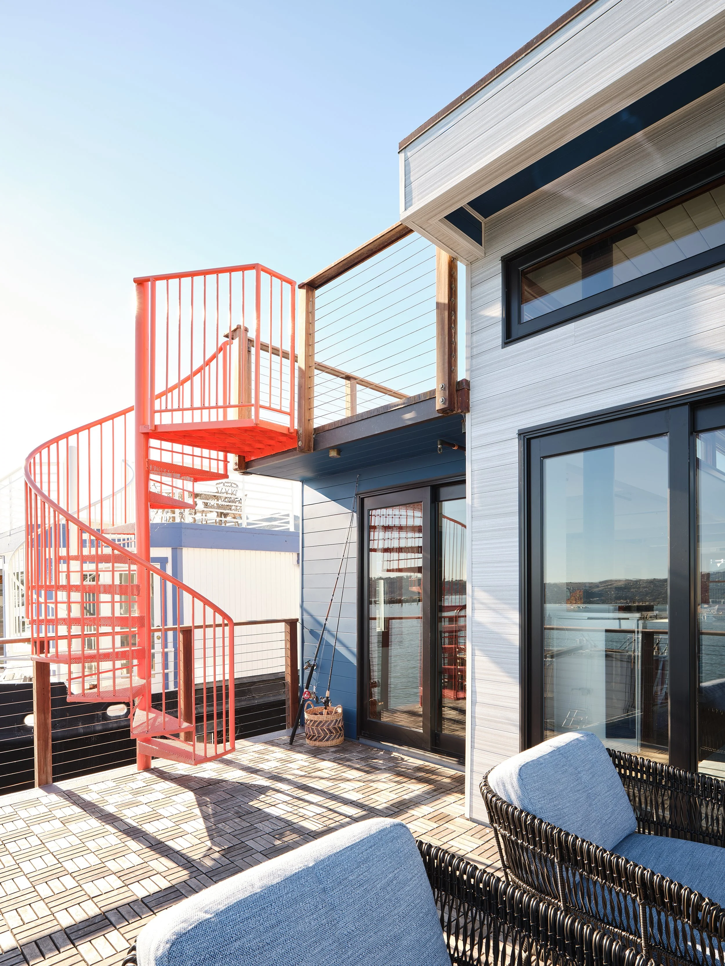 View of a rooftop outdoor patio with wooden flooring, black-framed glass doors, patio chairs, and a red spiral staircase leading to a balcony.