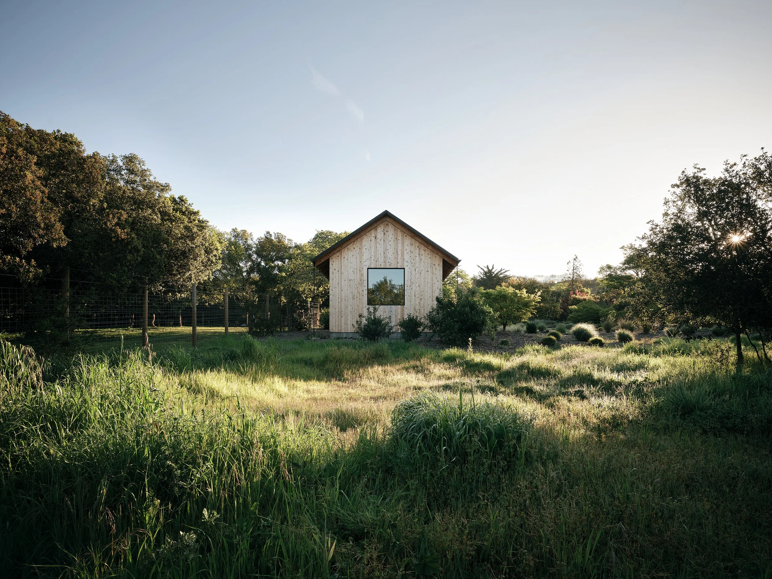 A small wooden house with a single window surrounded by lush green grass and trees, with the sun setting in the background.