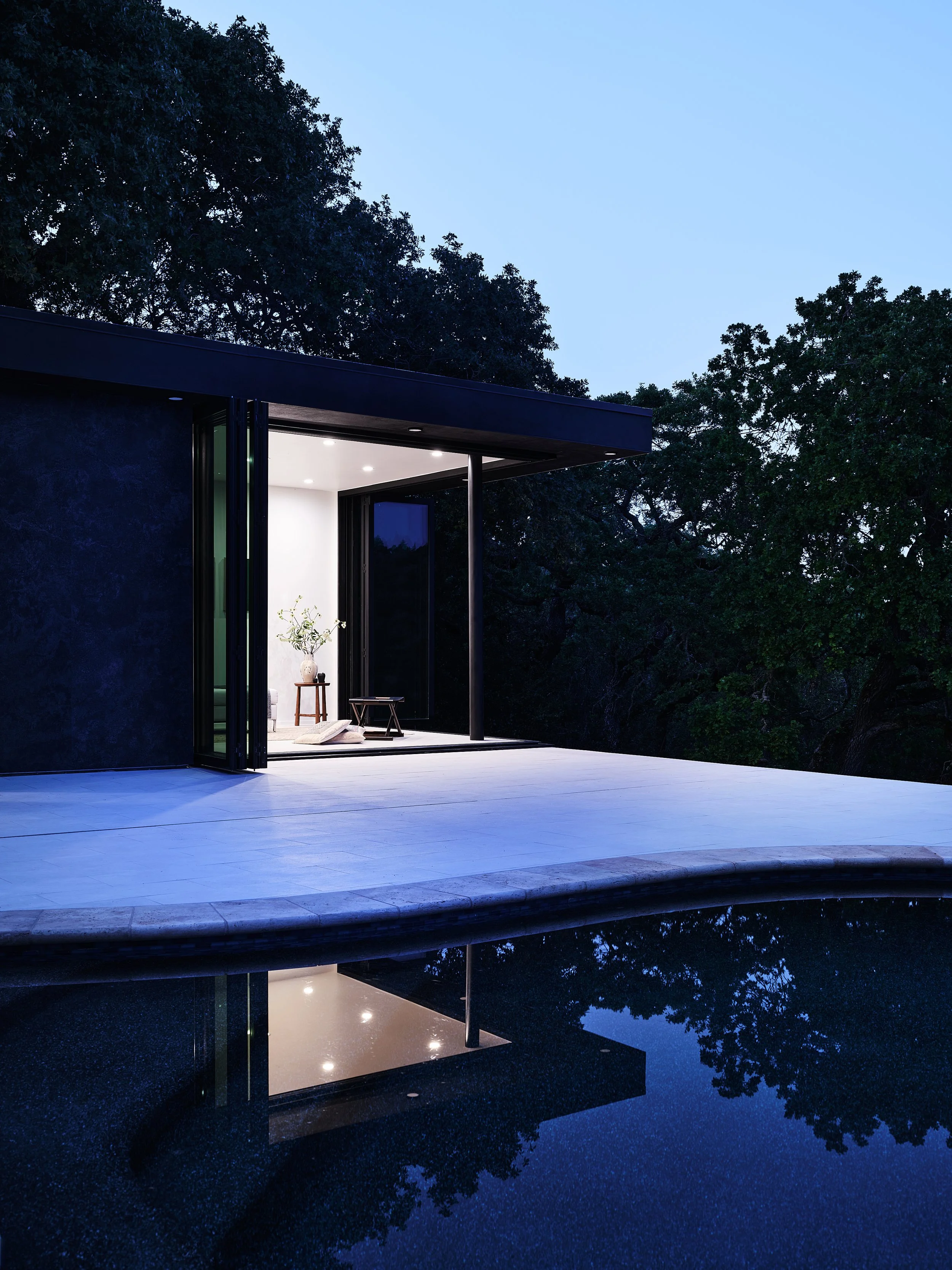 Modern house with an open sliding glass door, illuminated interior, and a reflective pool in the foreground surrounded by trees at dusk.