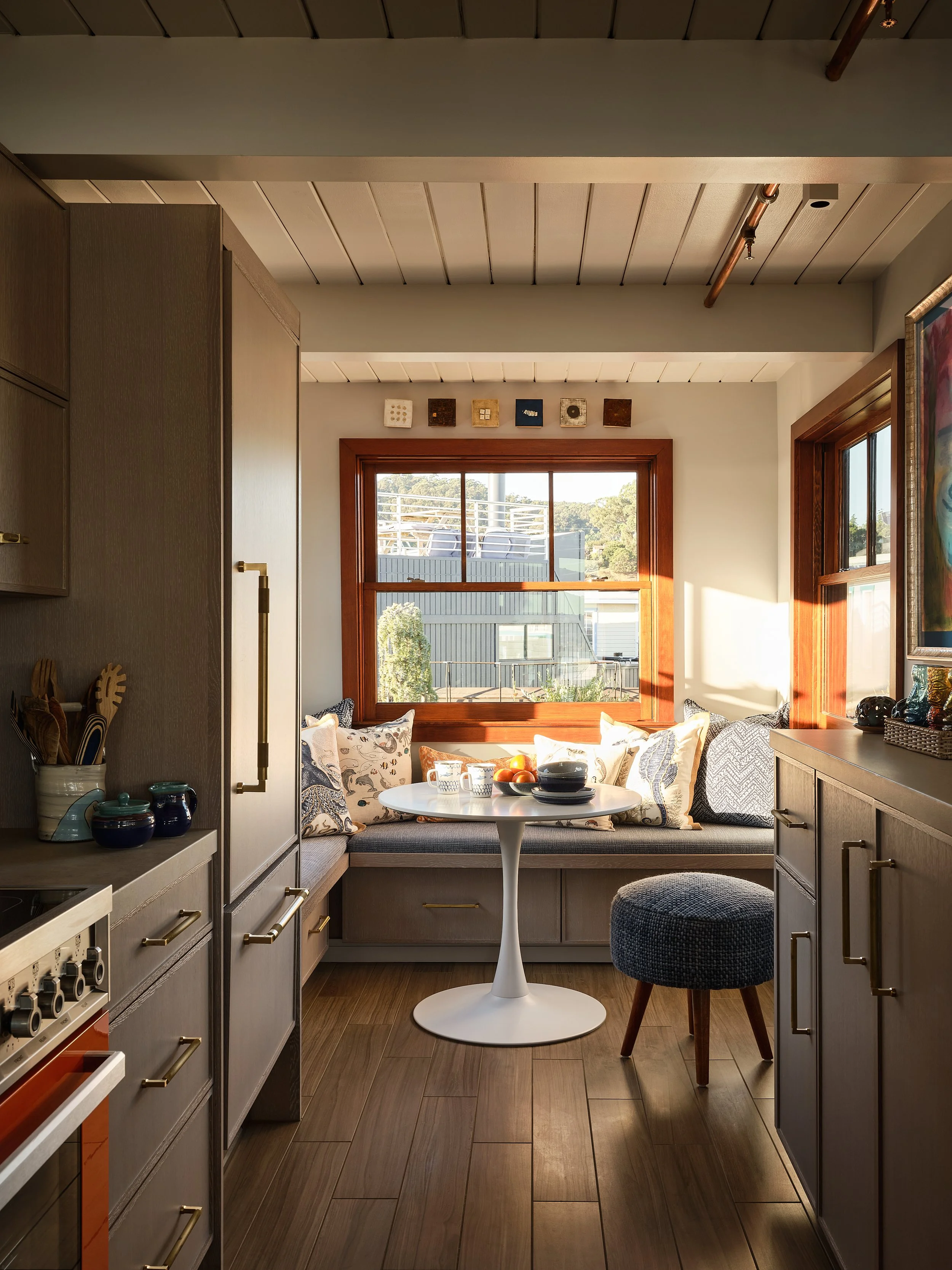Cozy kitchen nook with a round white table, cushions on a bench by the window, sunlight streaming through the wooden-framed window, and a small stool.