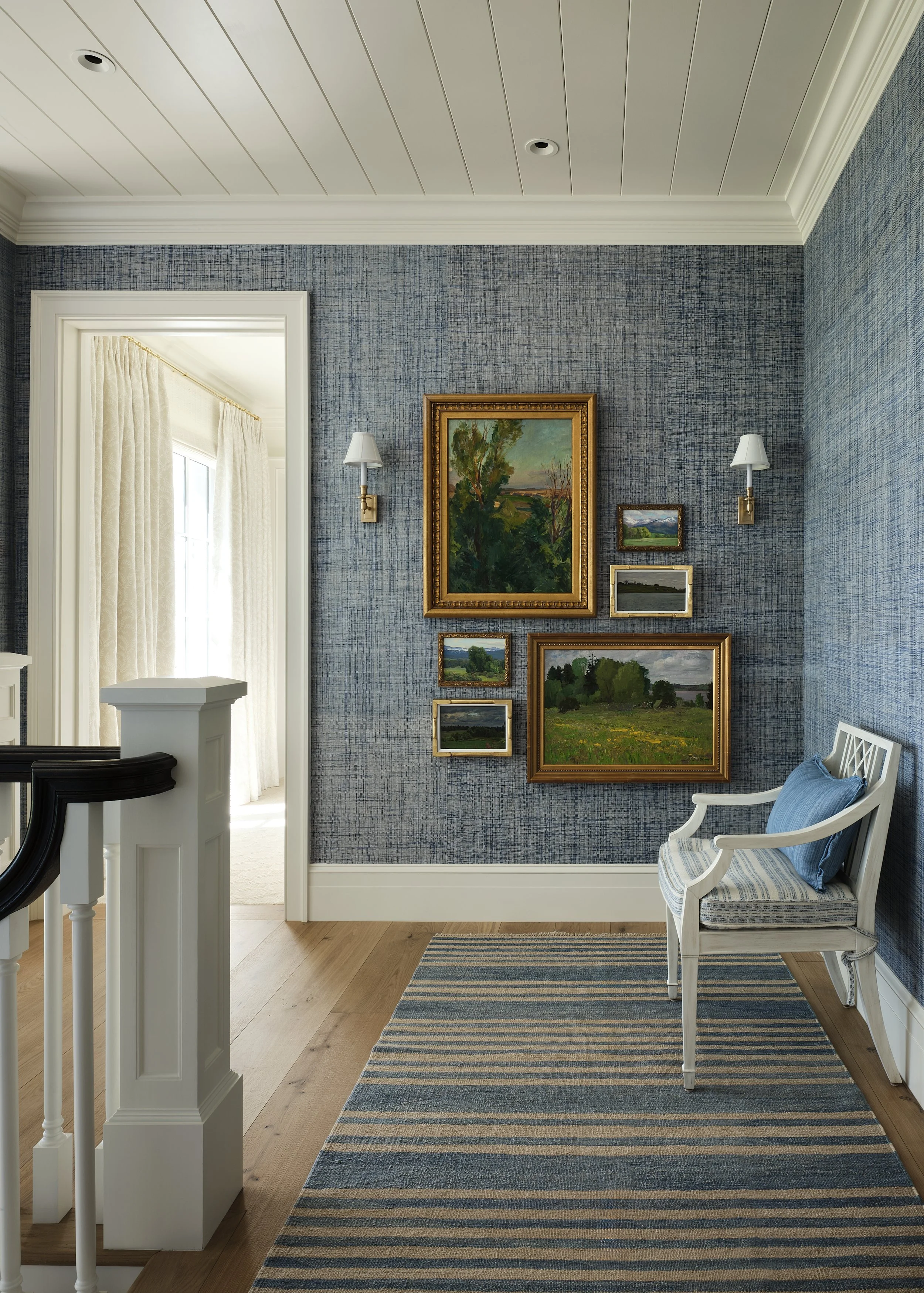 A cozy corner of a living room with blue textured wallpaper, a white wooden chair with a striped cushion and blue pillow, a striped rug, and a gallery wall of framed landscape paintings. Small wall sconces are on each side of the gallery.