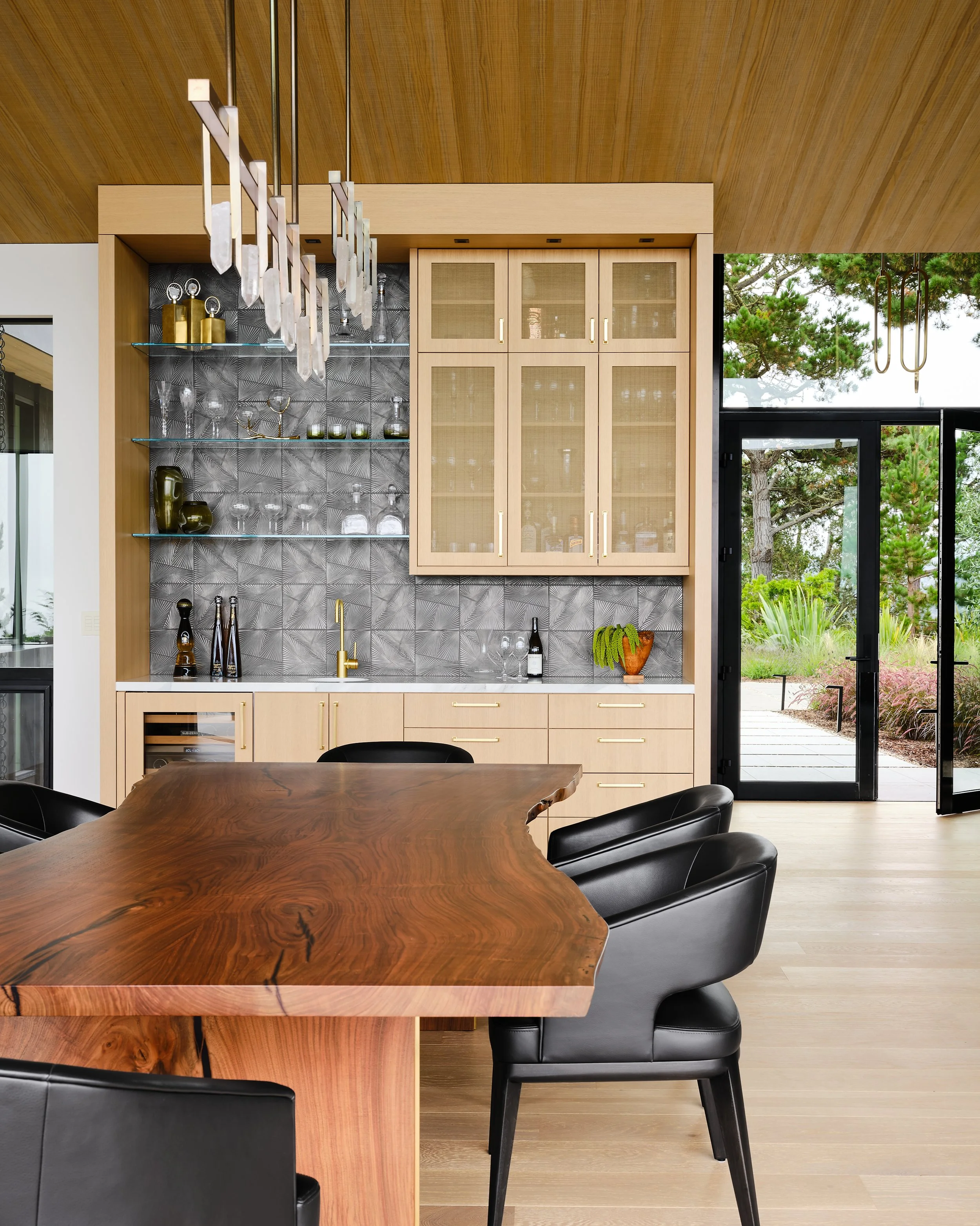 Modern dining area with a large wooden table, black leather chairs, a built-in cabinet with glass doors, and floor-to-ceiling windows showing outdoor greenery.