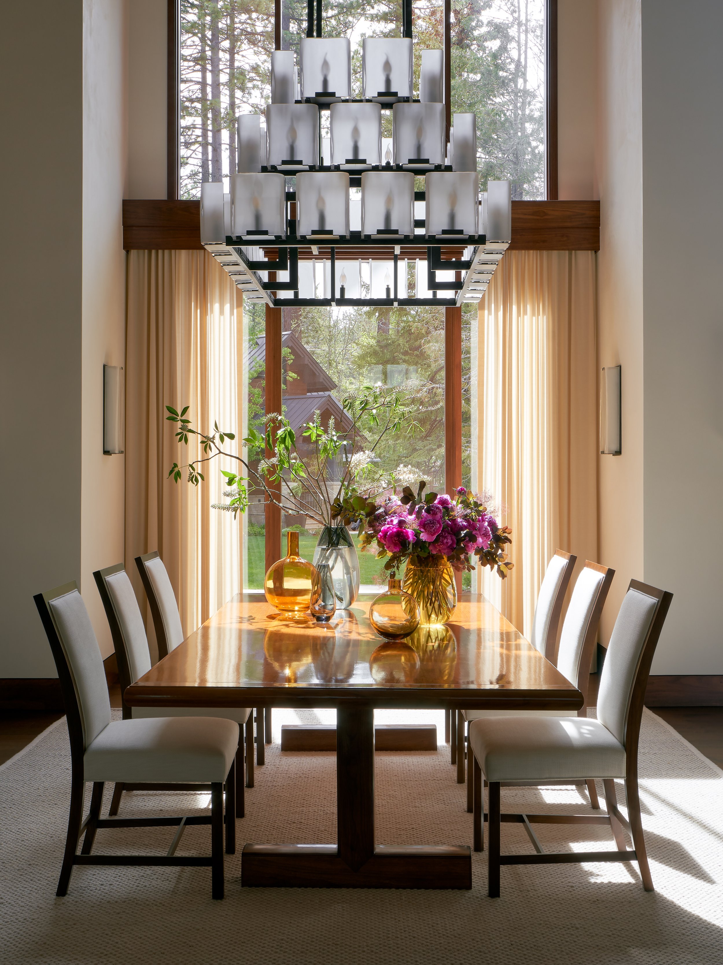 Dining room with a wooden table, six white chairs, a large window with curtains, a chandelier overhead, and a floral centerpiece with vases of flowers and foliage.