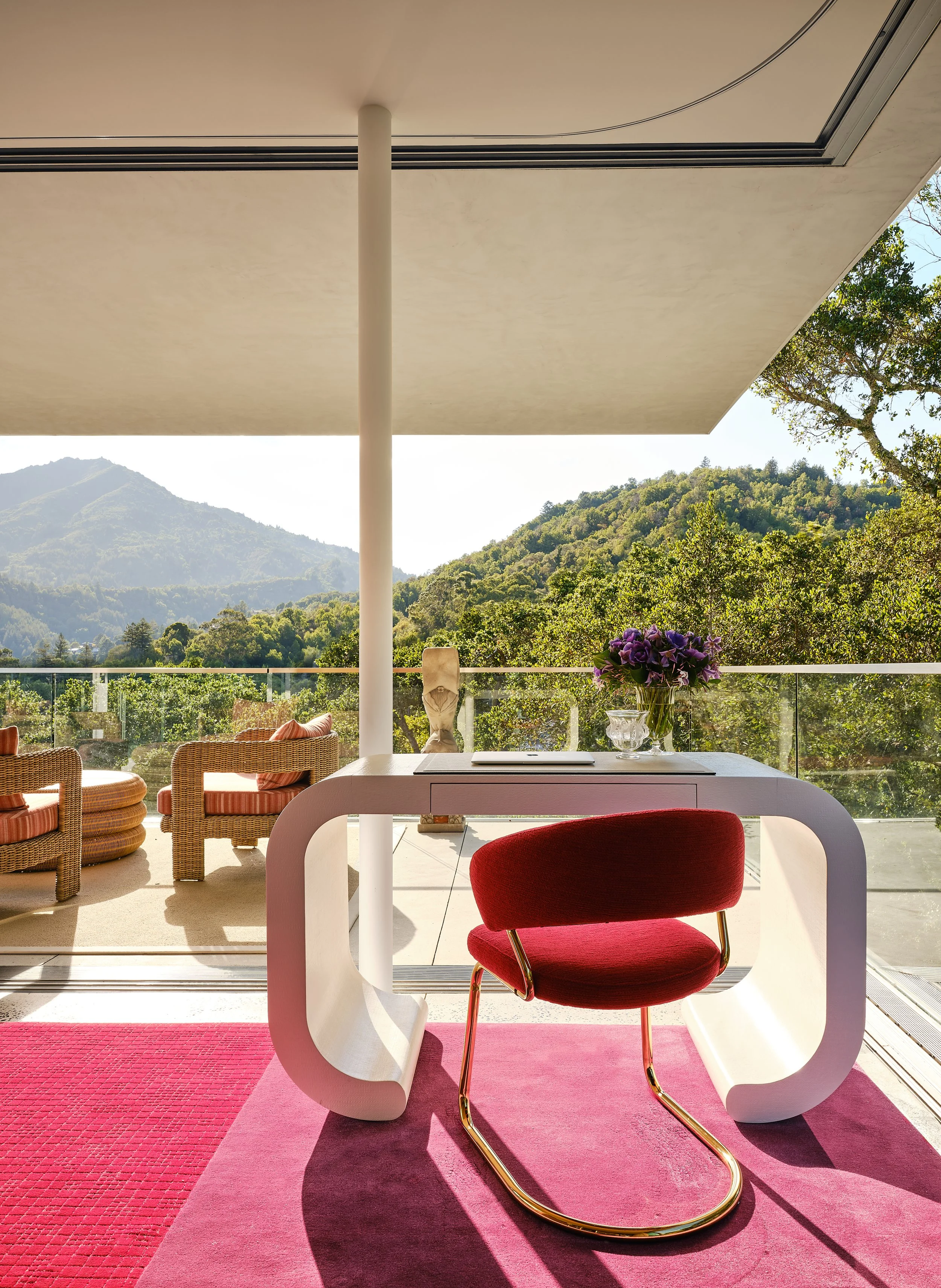 Modern indoor workspace with a white desk and red chair, overlooking a mountain view with greenery and trees outside, with wicker chairs on a balcony in the background.