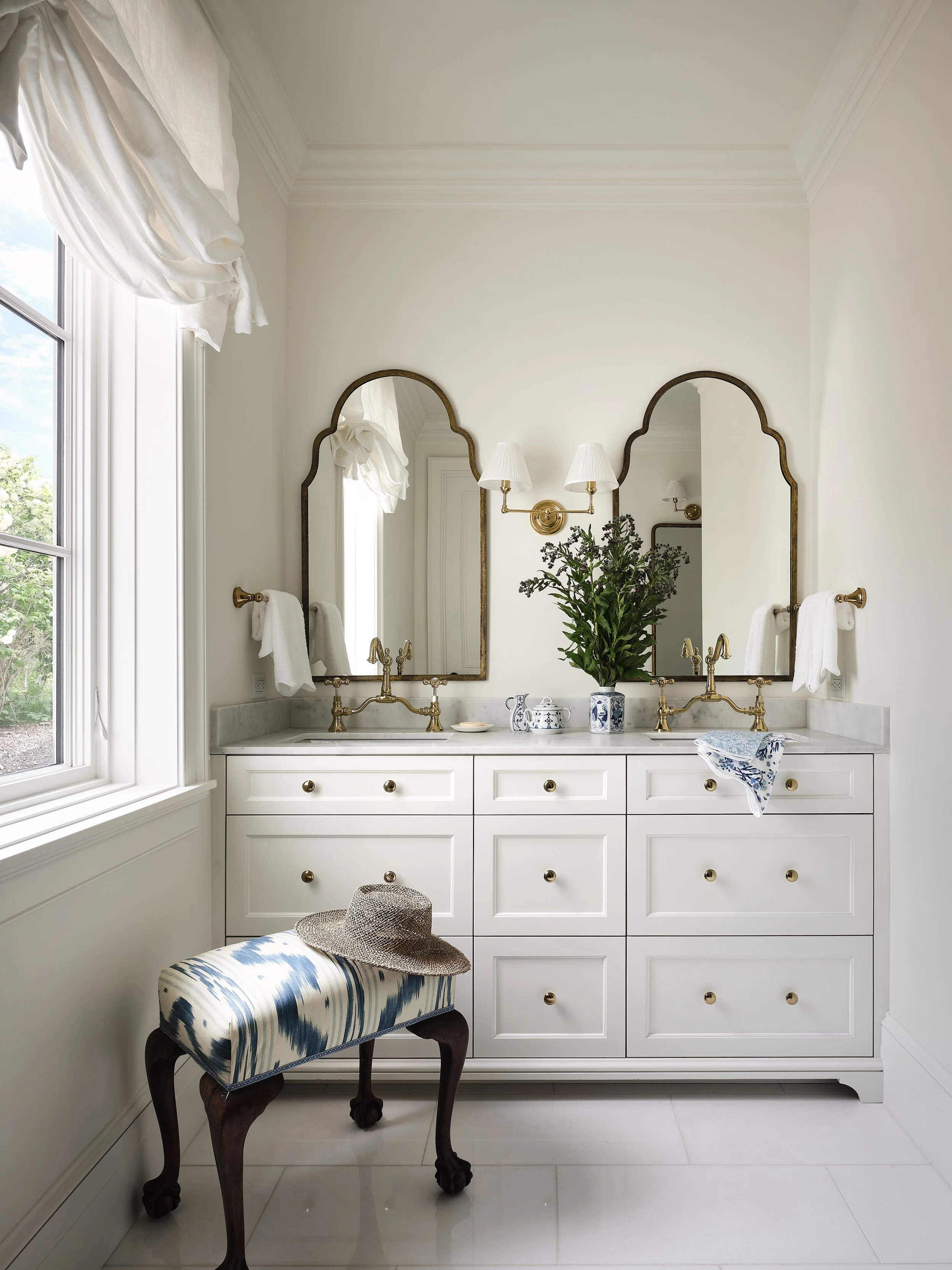 Bright bathroom with white cabinetry, two gold faucets, and two tall mirrors with scalloped edges. There are white towels on gold towel bars, a potted plant between the mirrors, and a blue and white floral dish towel hanging from a drawer. A window w