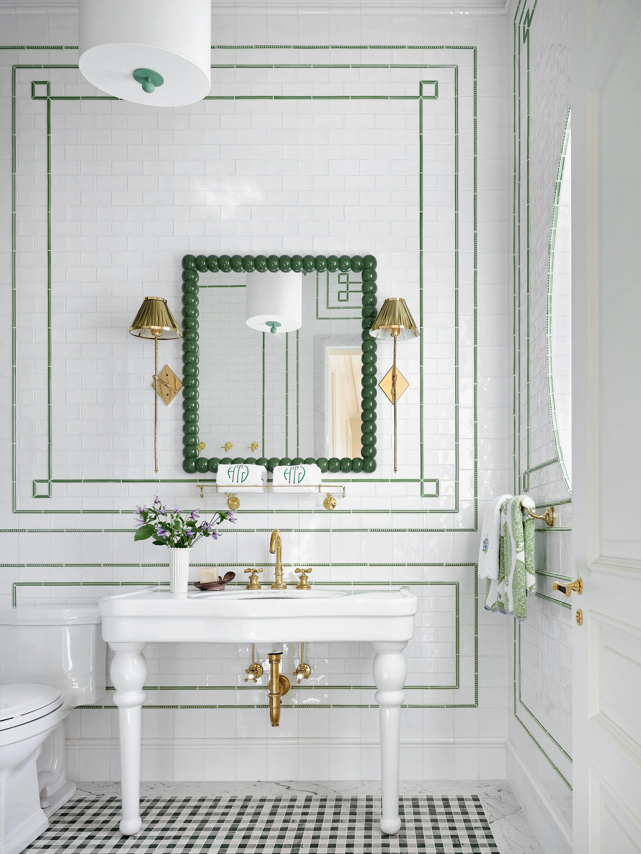 A white bathroom with green decorative tiles, a large mirror with a frame of green beads, wall-mounted gold lamps with patterned shades, a white vintage-style sink with gold fixtures, and a flower vase with purple flowers on the sink.