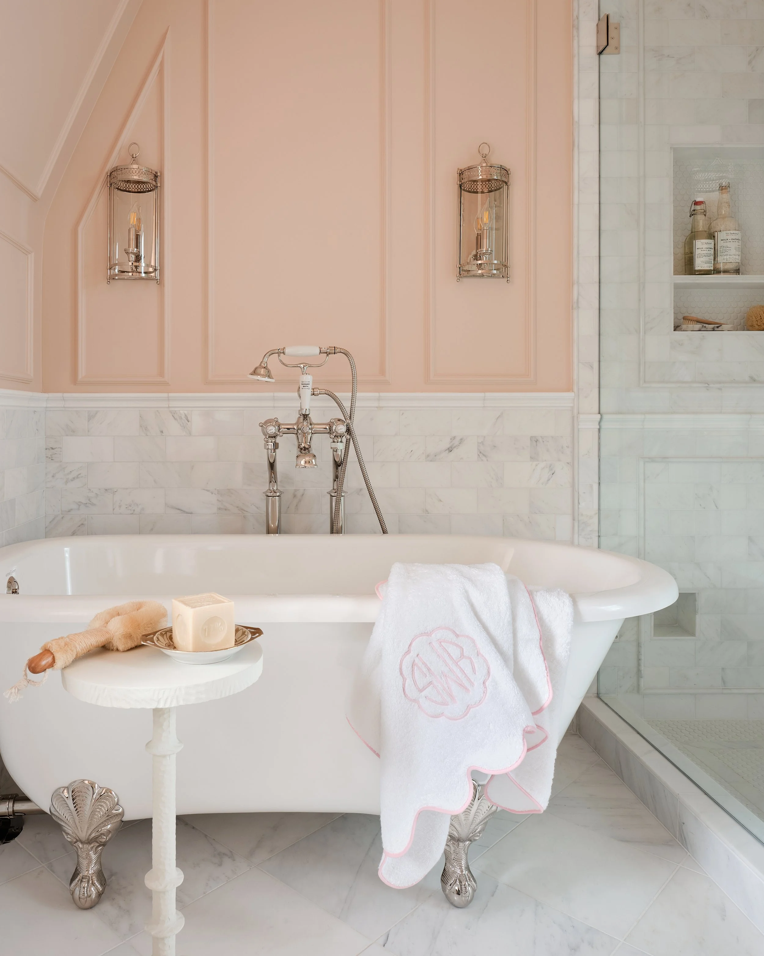 Bathroom with a vintage white clawfoot bathtub, a towel with monogram, and a small side table with a soap and loofah, all in a luxurious setting with pink and marble accents.