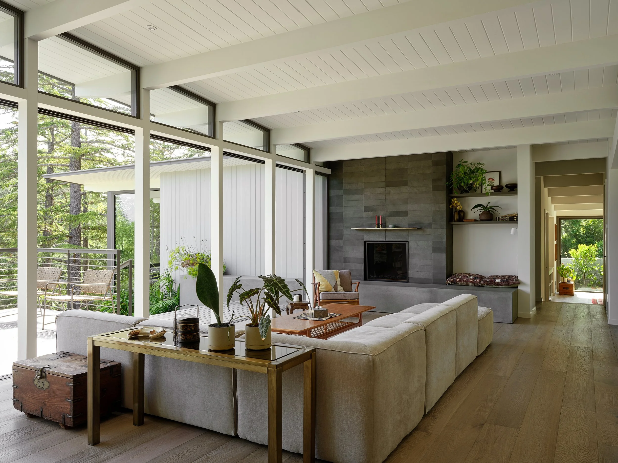 Living room with large windows, modern fireplace, beige sofa, wooden coffee table, and decor plants.