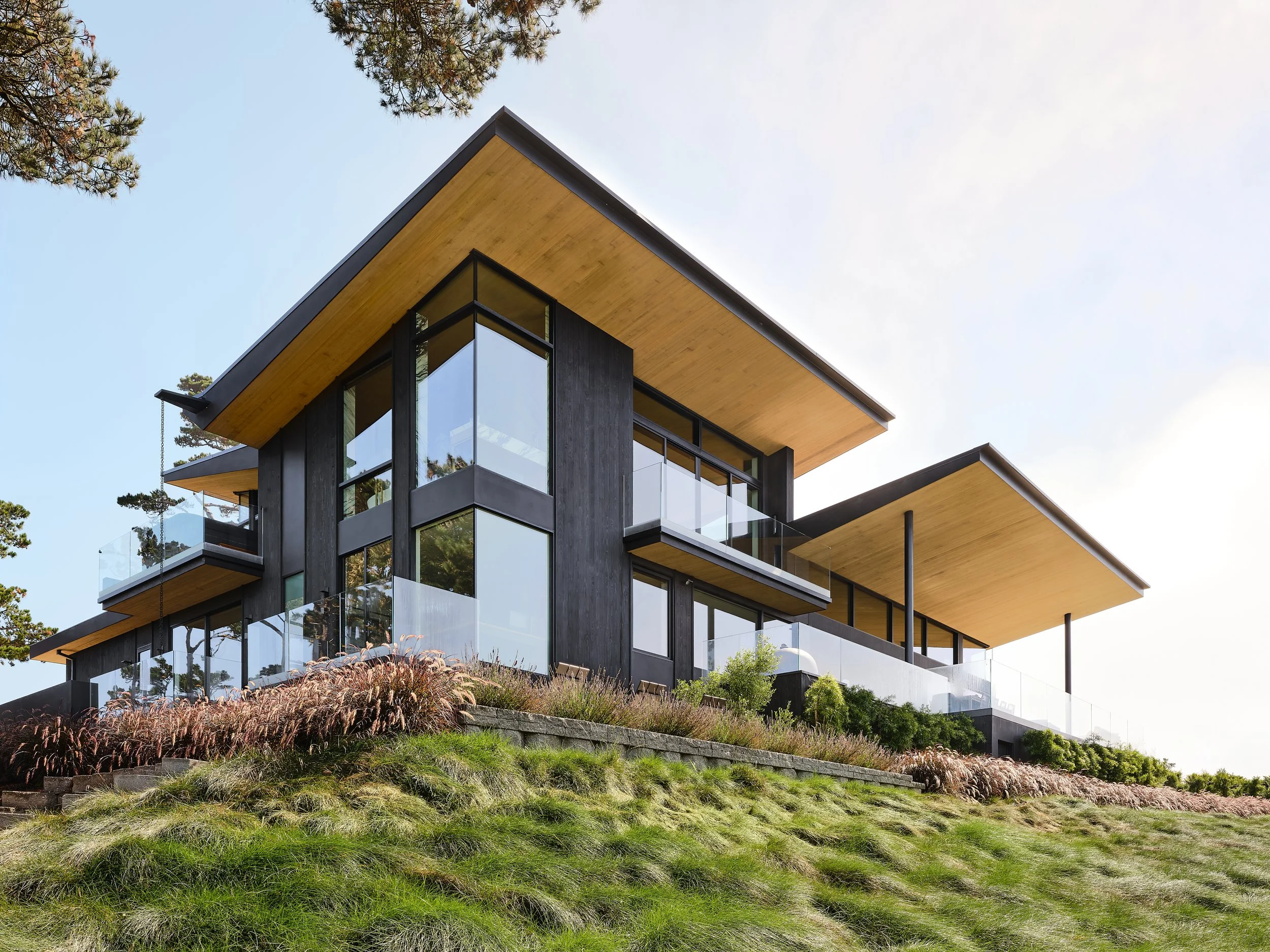 Modern multi-level house with large glass windows, black exterior walls, and wooden overhangs on a grassy hillside.
